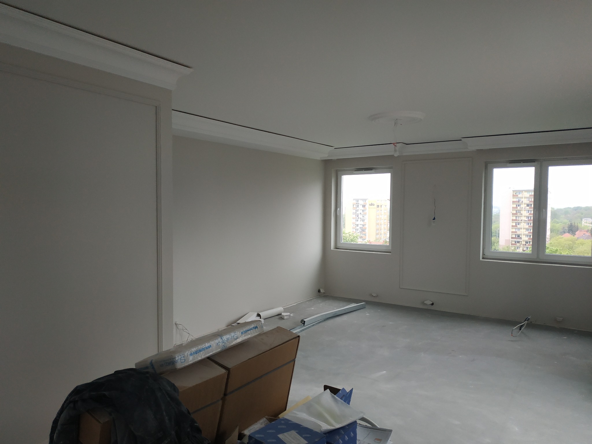 Pokój w trakcie remontu: pomalowane ściany na biało, listwy dekoracyjne przy suficie, szpachlowana podłoga, widoczne materiały budowlane i kartony.