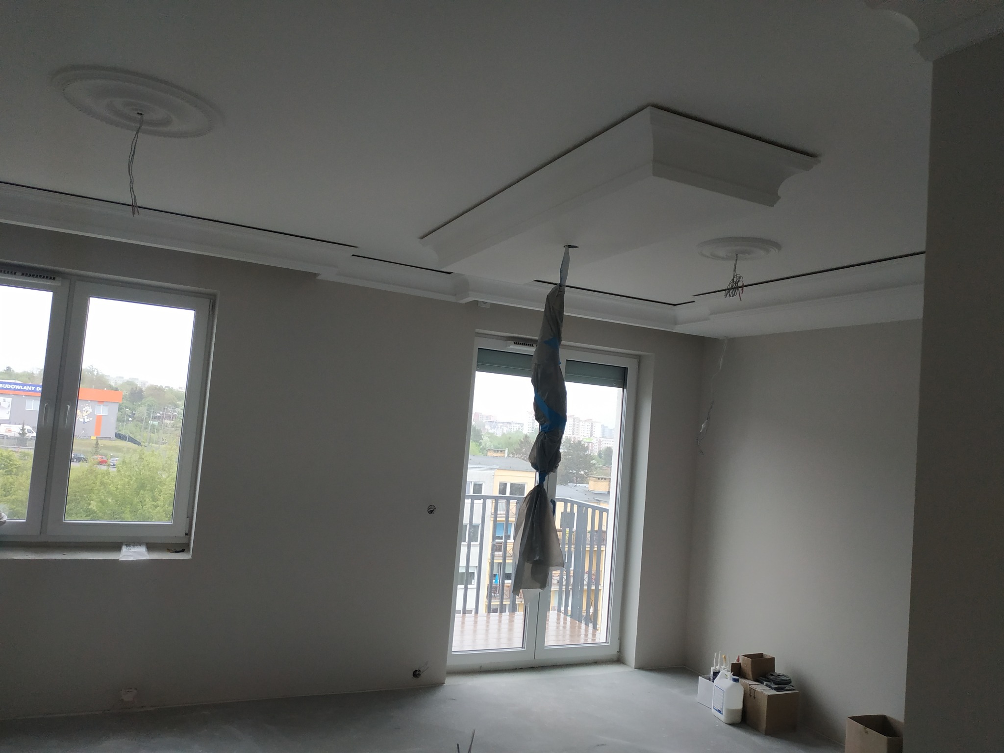 Wnętrze w trakcie remontu: surowa, szpachlowana ściana, dekoracyjne listwy przysufitowe i rozety, zabezpieczone folią drzwi balkonowe, widoczne puszki i kartony z materiałami budowlanymi.