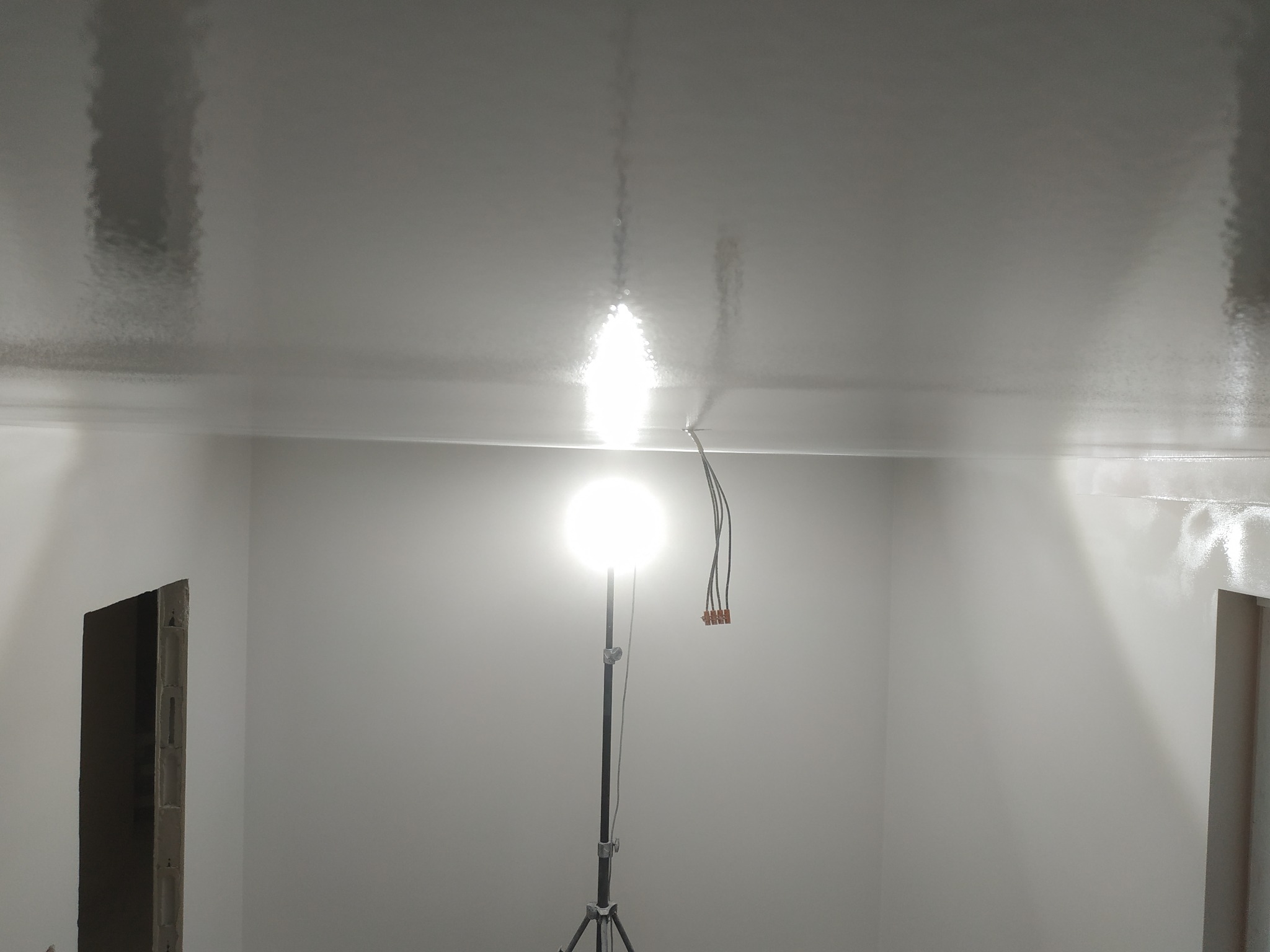 Świeżo pomalowany, błyszczący sufit w pokoju, oświetlony lampą budowlaną na statywie, widoczne kable lampy, przygotowane do dalszych prac wykończeniowych, stan surowy w narożnikach.