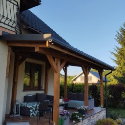 Tomasz Rychlik - Zadaszony taras z drewnianą konstrukcją, wyposażony w meble wypoczynkowe i dekoracje kwiatowe, z widokiem na ogród i sąsiedni dom.