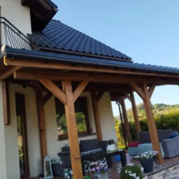 Zadaszony taras z drewnianą konstrukcją, meblami wypoczynkowymi i kwiatami w donicach, przylegający do domu z ciemnym dachem.