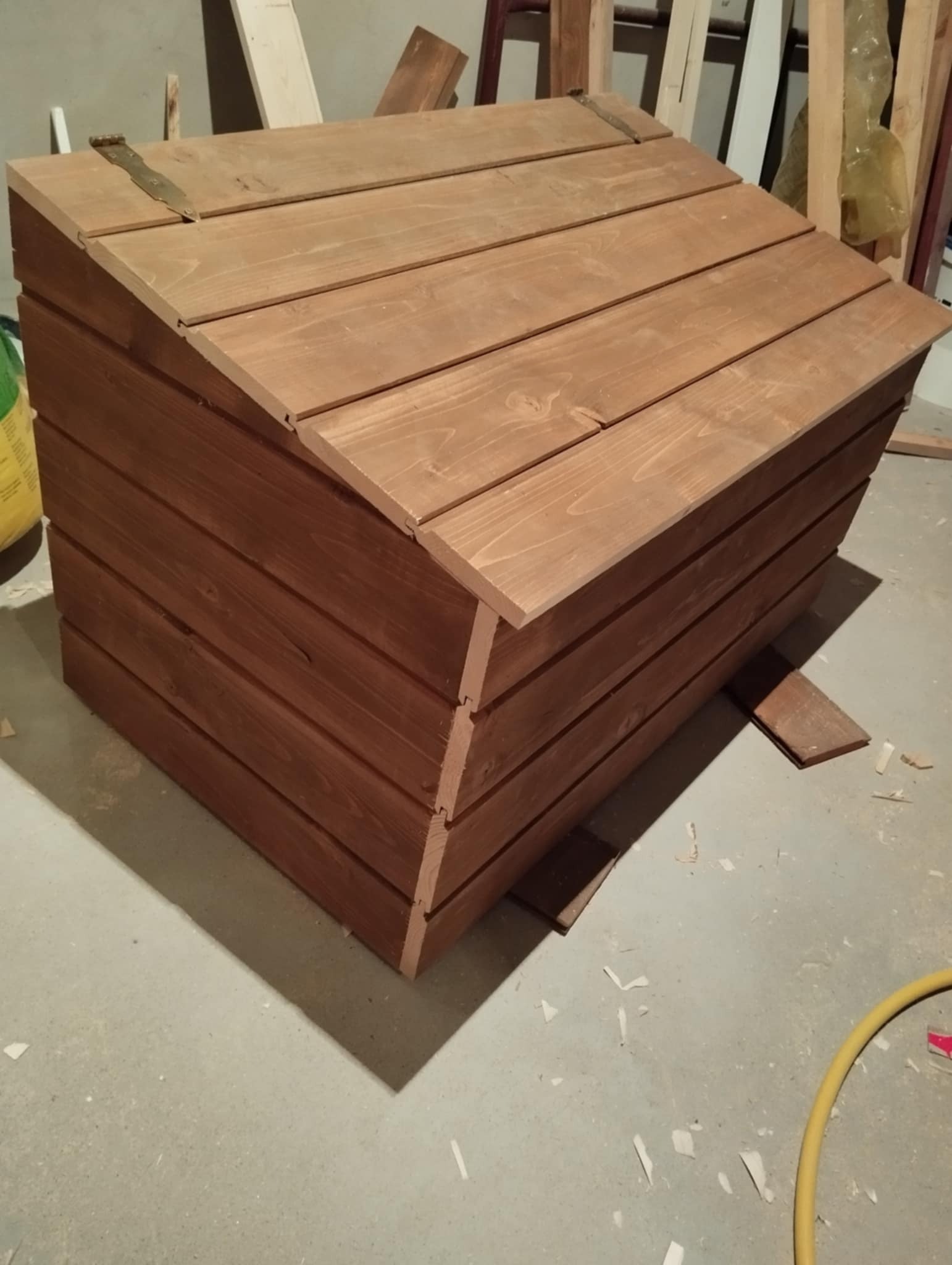 Drewniana skrzynia na narzędzia lub zabawki, wykonana z impregnowanych desek, zamykana na metalowe zawiasy, widoczna na tle surowego garażu z resztkami drewna na podłodze.