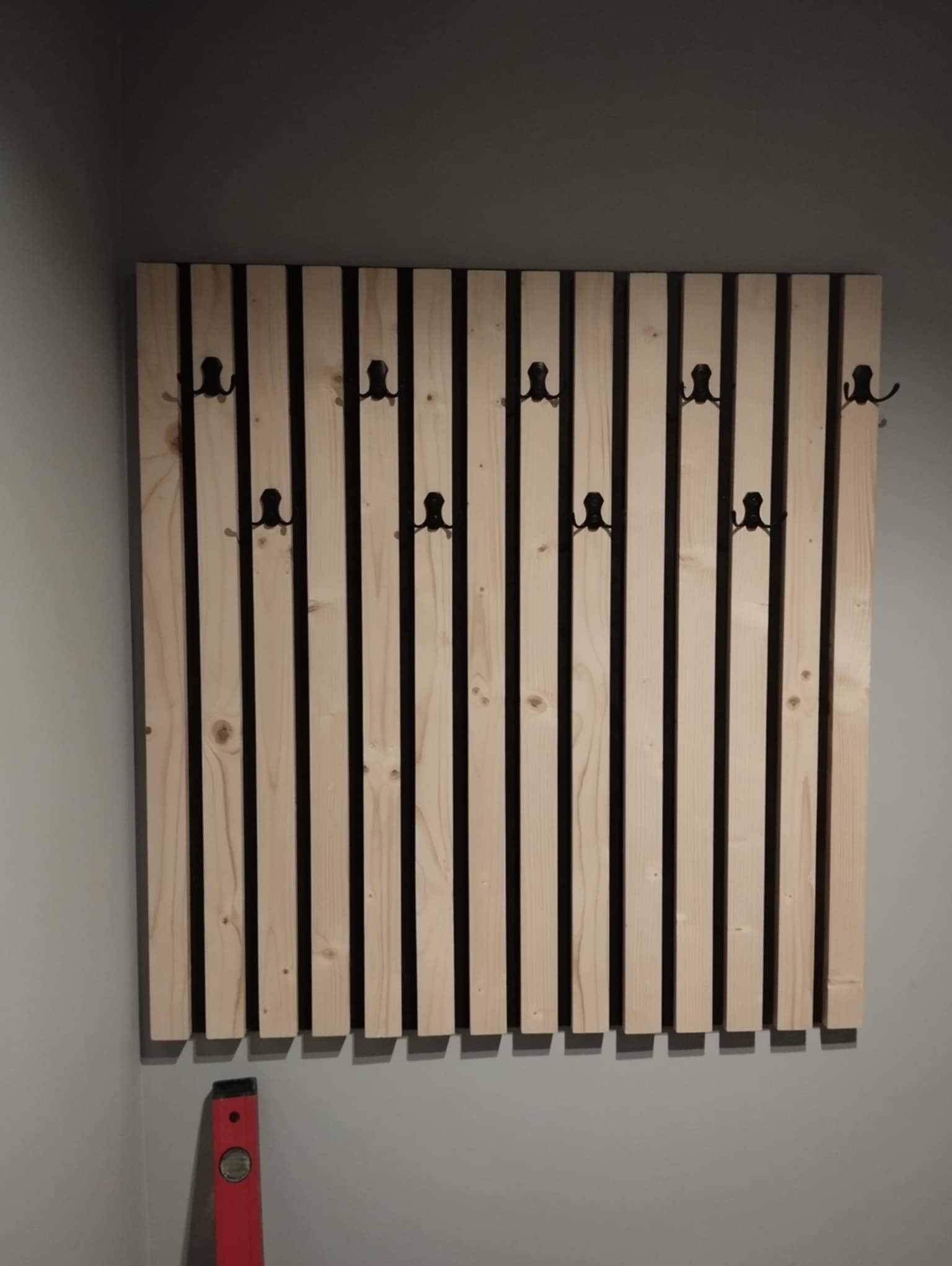 Drewniany panel ścienny z czarnymi haczykami, zamontowany w Świnnej, z użyciem poziomicy dla precyzyjnego montażu.
