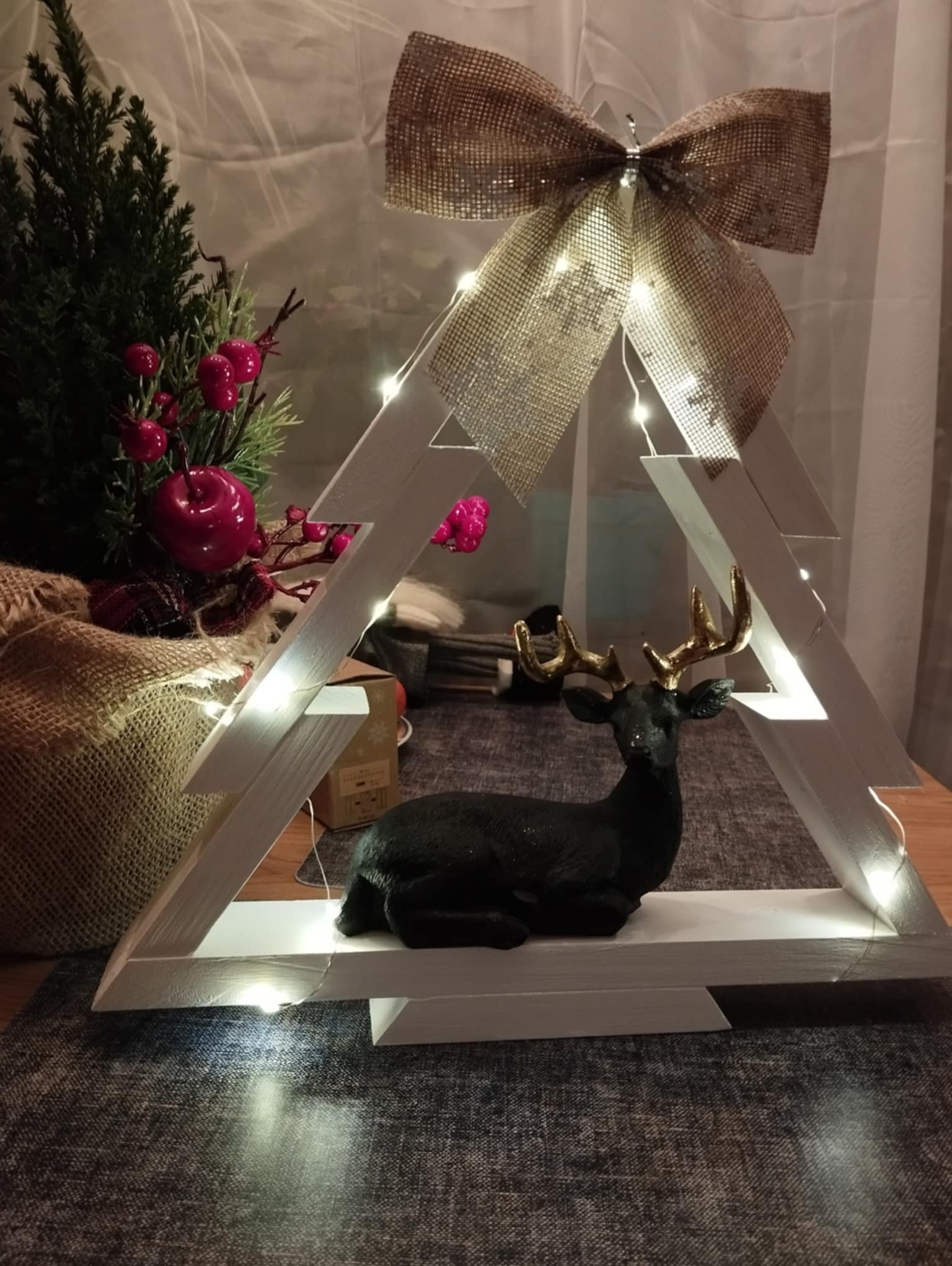 Dekoracja świąteczna: ręcznie robiona drewniana choinka z oświetleniem LED, ozdobiona jutowym kokardą i czarnym reniferem z złotymi rogami, obok mała choinka z czerwonymi jabłkami.