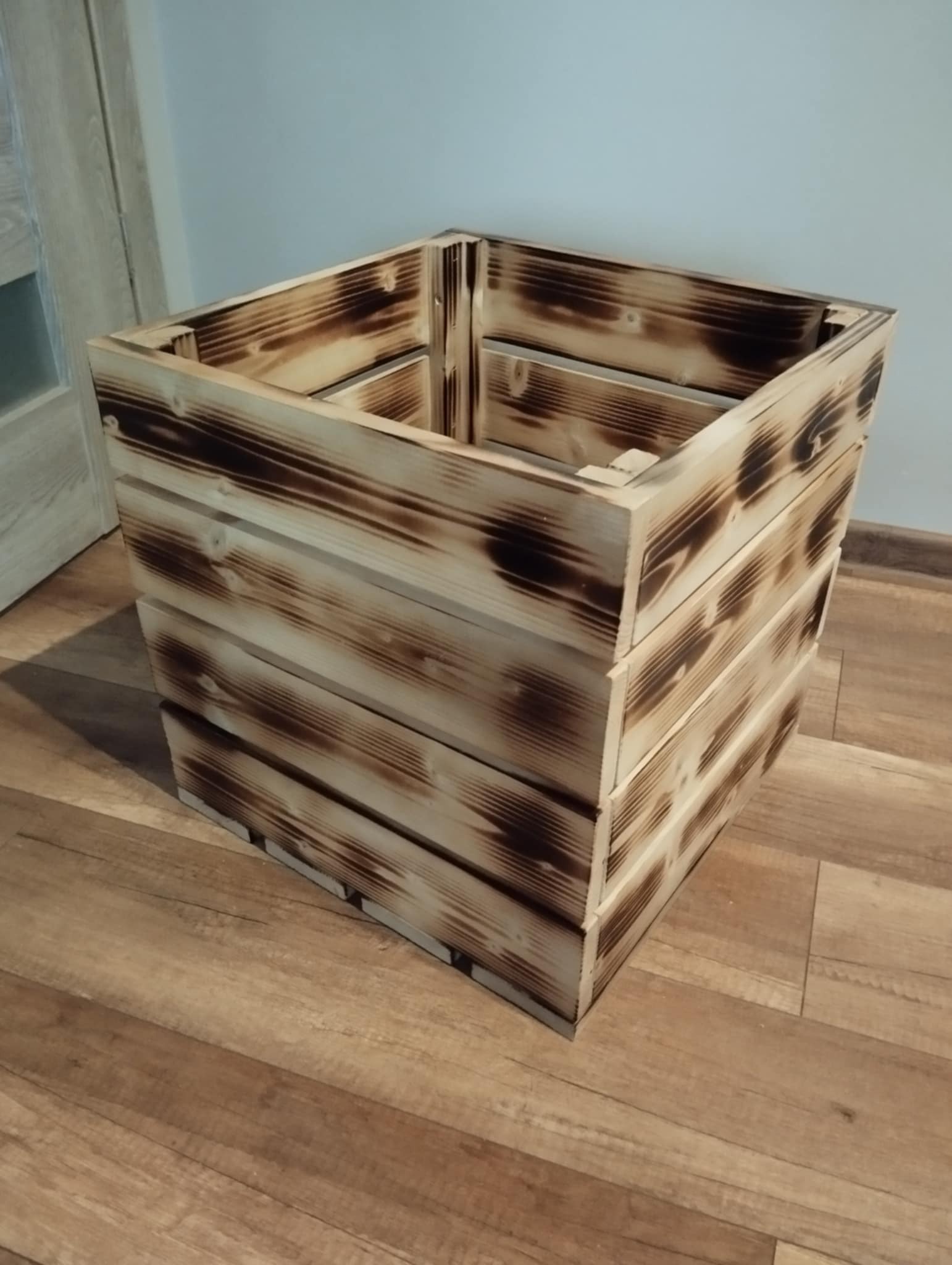 Kwadratowa, drewniana skrzynia z efektem opalania, widoczna na drewnianej podłodze w jasnym pomieszczeniu.