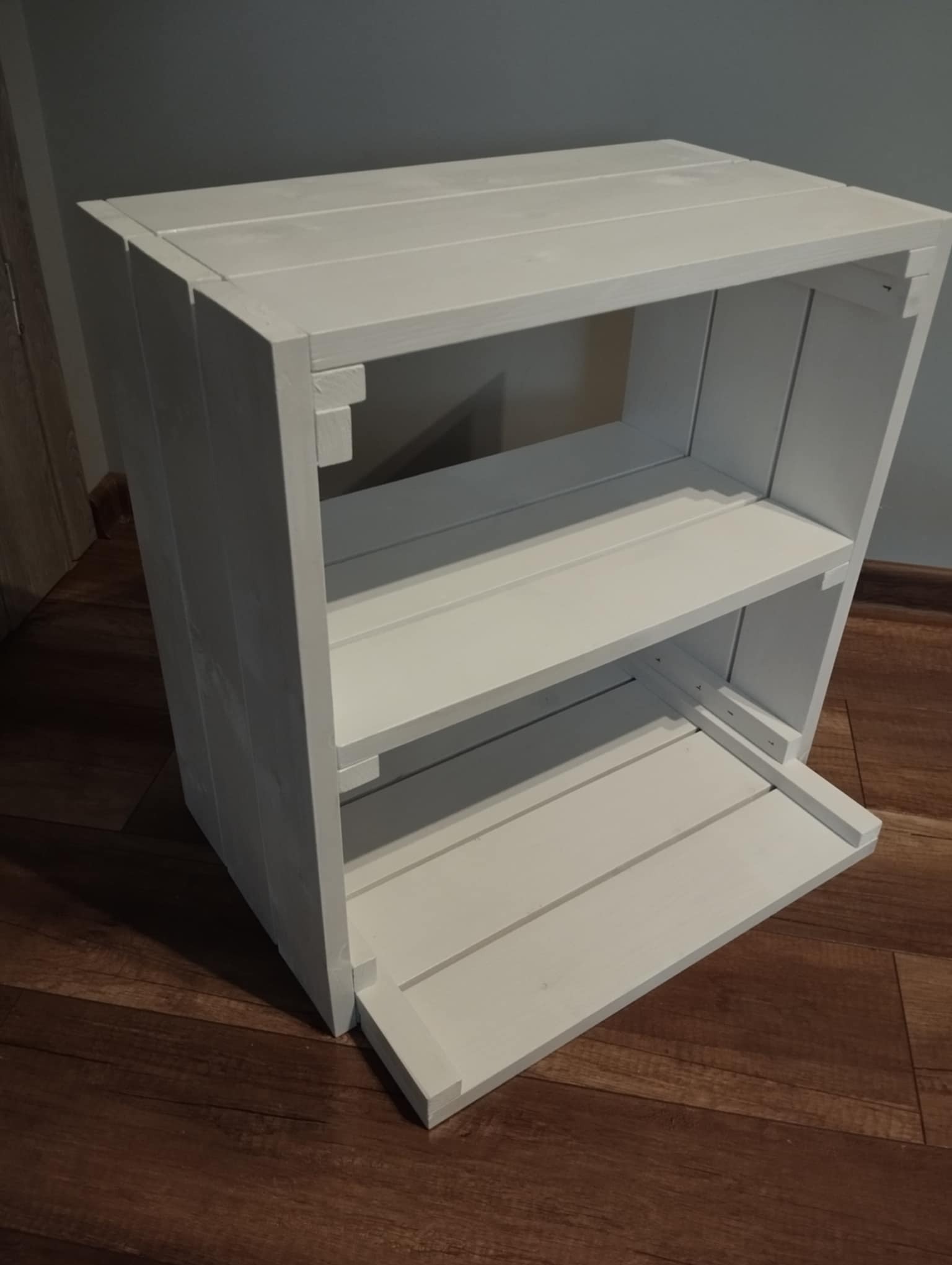 Biały, drewniany regał z trzema półkami, wykonany z desek, stojący na brązowej podłodze, na tle szarej ściany.