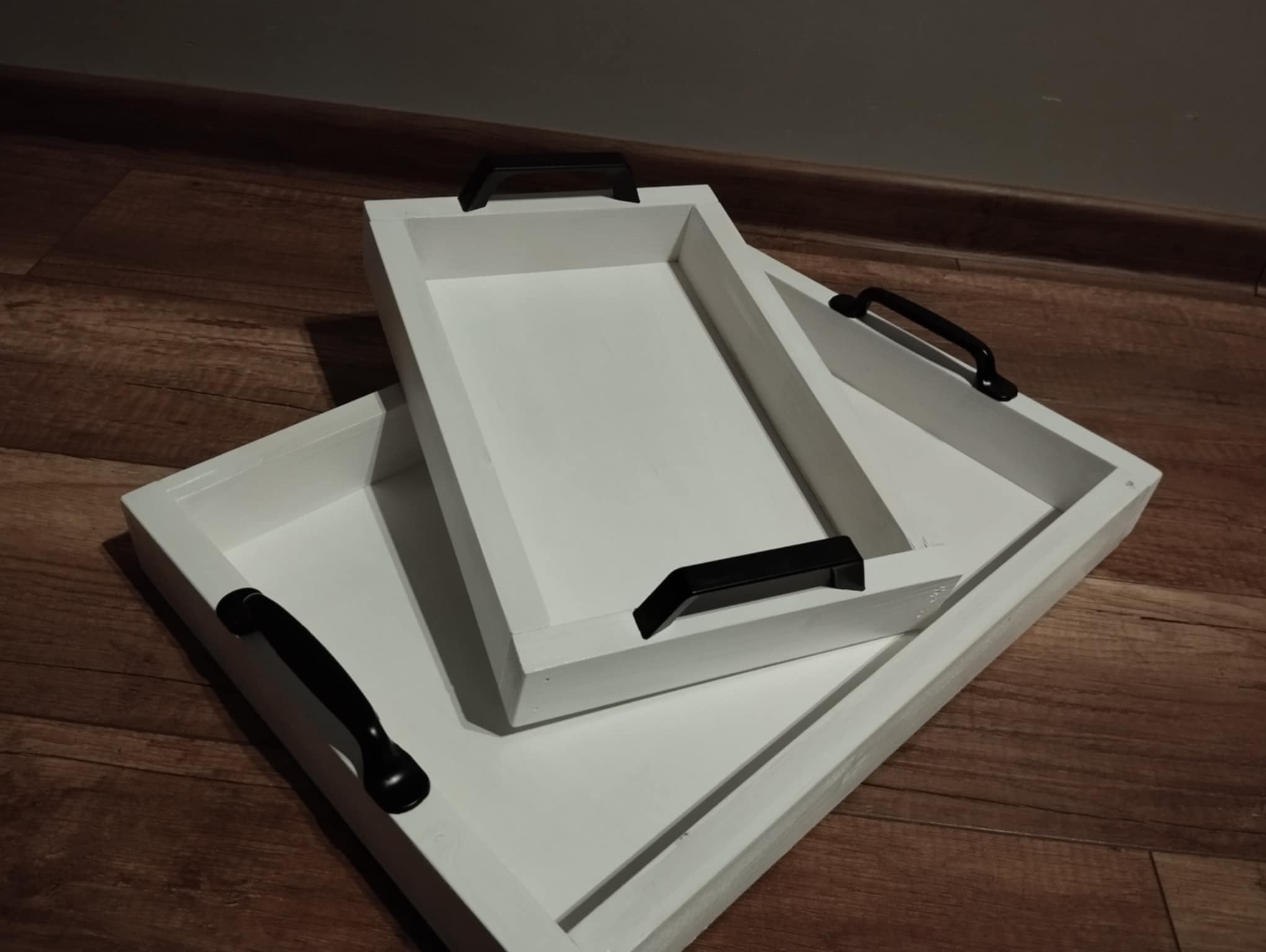 Trzy białe, kwadratowe tace dekoracyjne z czarnymi uchwytami, ułożone jedna na drugiej na drewnianej podłodze. Tace mają proste, minimalistyczne wykończenie.