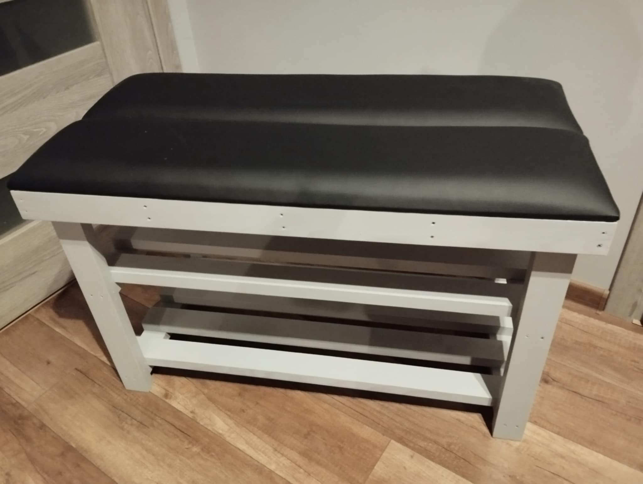 Biała, drewniana ławka z czarnym, tapicerowanym siedziskiem i półkami na buty, ustawiona na panelach podłogowych.