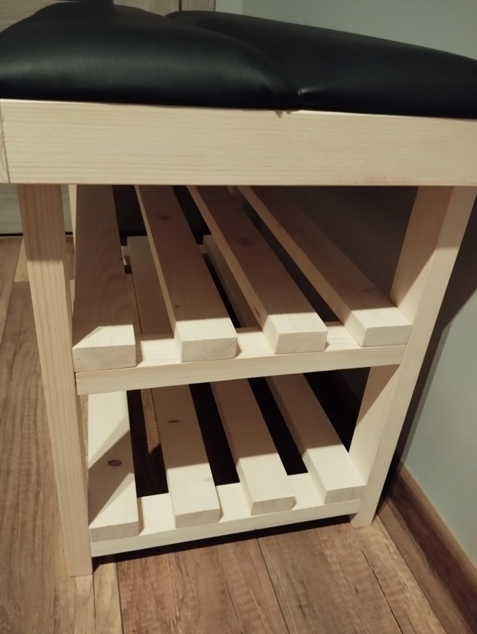 Drewniany regał na buty z czarnym siedziskiem z ekoskóry, widok z boku na trzy półki z listew, jasne drewno, podłoga imitująca drewno.