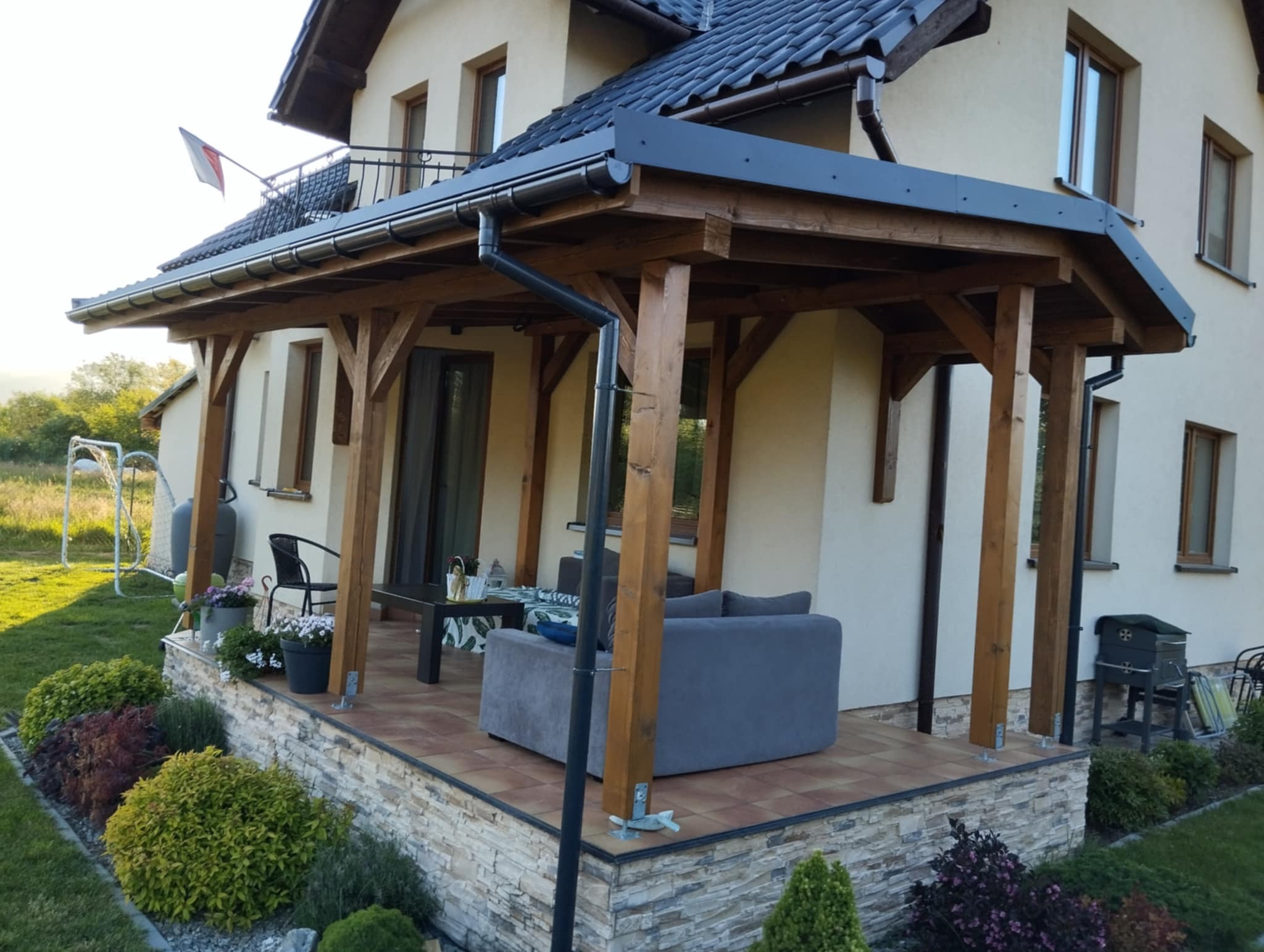 Zadaszony taras drewniany z szarą sofą i stolikiem kawowym, otoczony ozdobnym kamieniem i zadbanym ogrodem, z widoczną rynną i elementami konstrukcyjnymi z drewna.