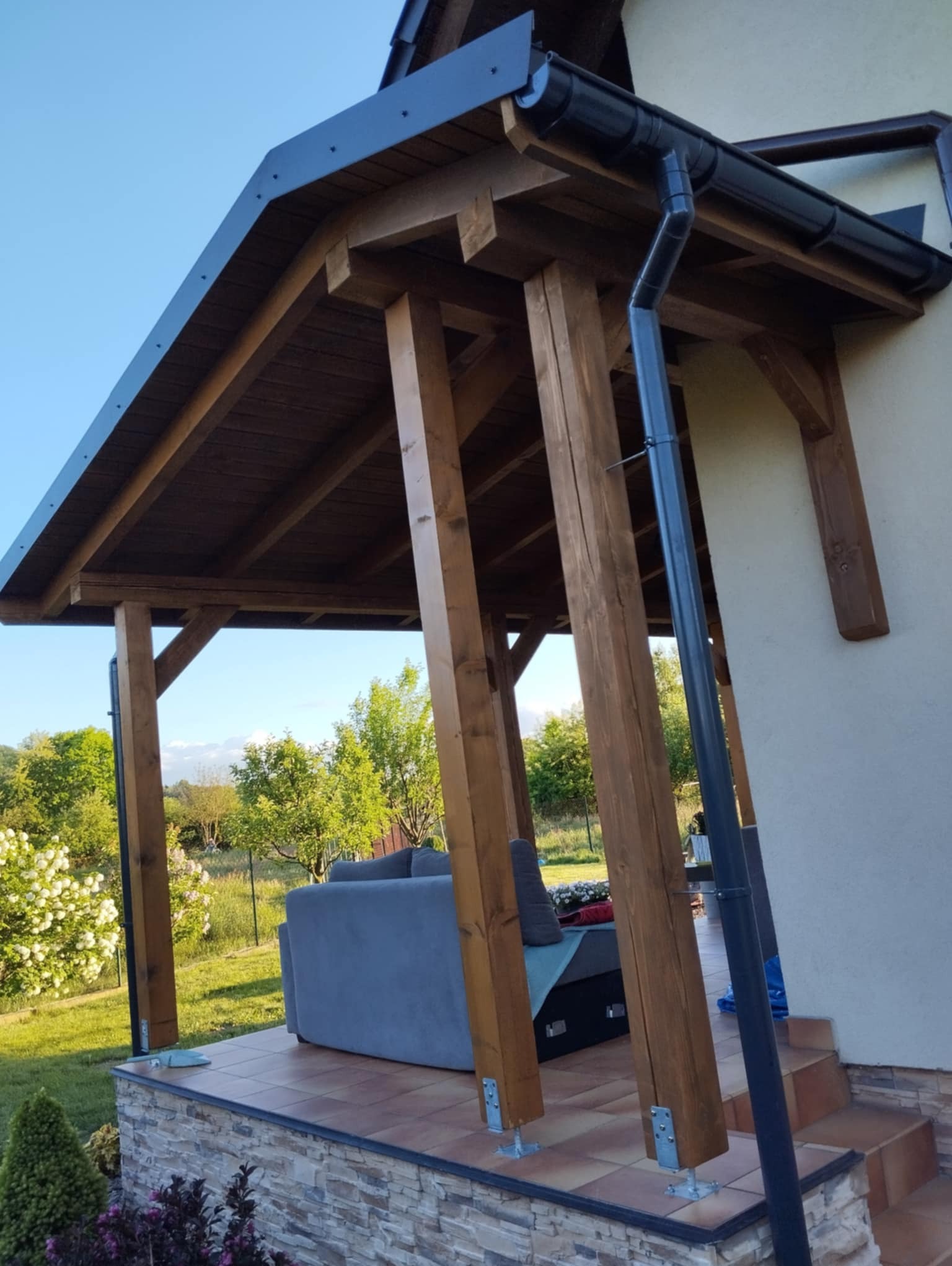 Zadaszony taras z drewnianą konstrukcją, szarym narożnikiem i kamienną podmurówką, widok z zewnątrz w słoneczny dzień.