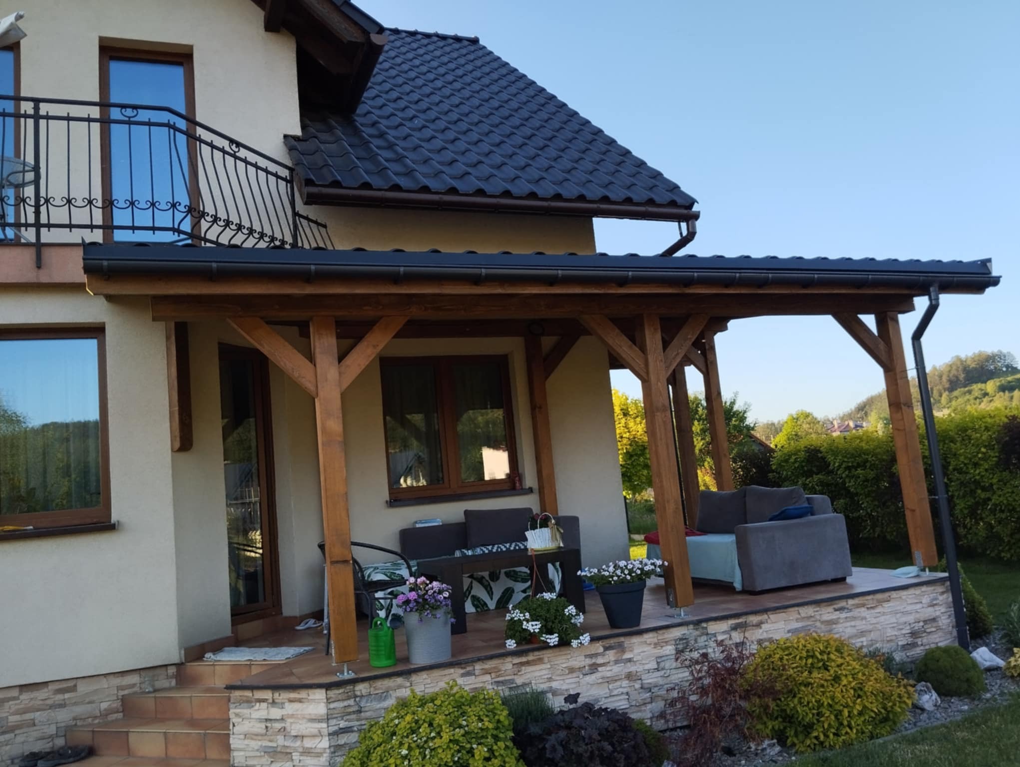 Zadaszony taras przy domu z drewnianą konstrukcją, meblami ogrodowymi i kwiatami w donicach, widok na zadbany ogród.