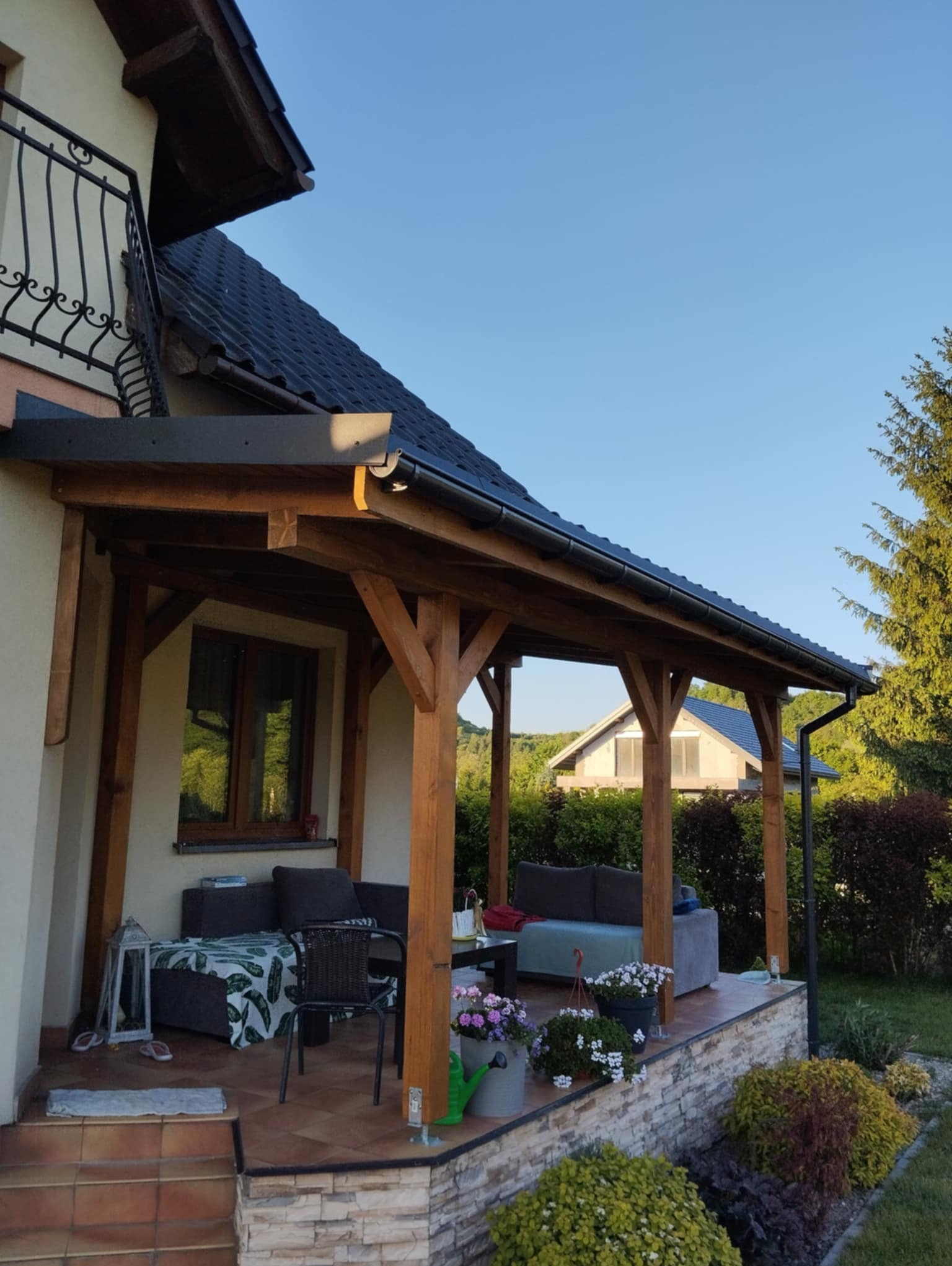 Zadaszony taras z drewnianą konstrukcją, wyposażony w meble wypoczynkowe i dekoracje kwiatowe, z widokiem na ogród i sąsiedni dom.