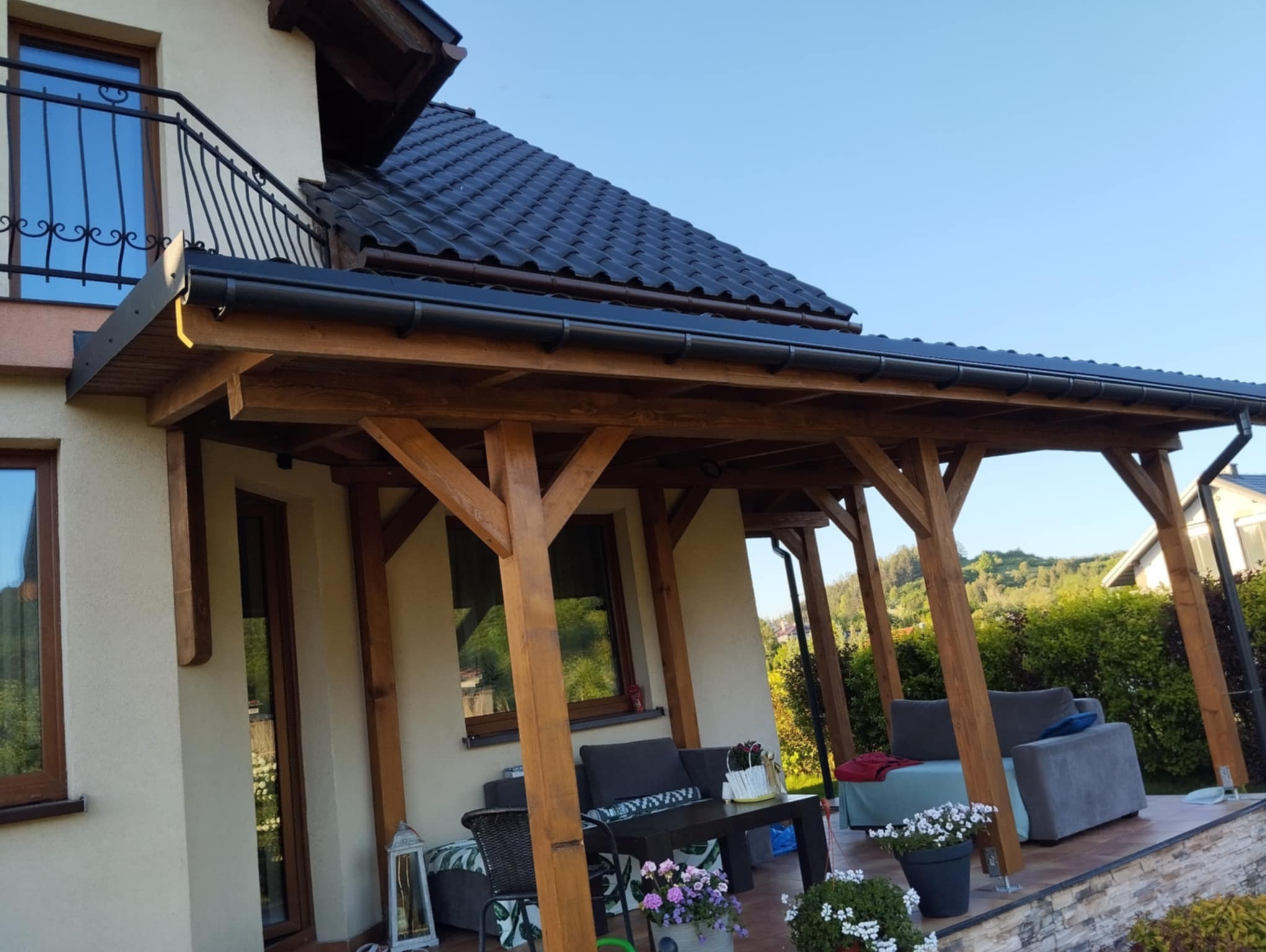 Zadaszony taras z drewnianą konstrukcją, meblami wypoczynkowymi i kwiatami w donicach, przylegający do domu z ciemnym dachem.
