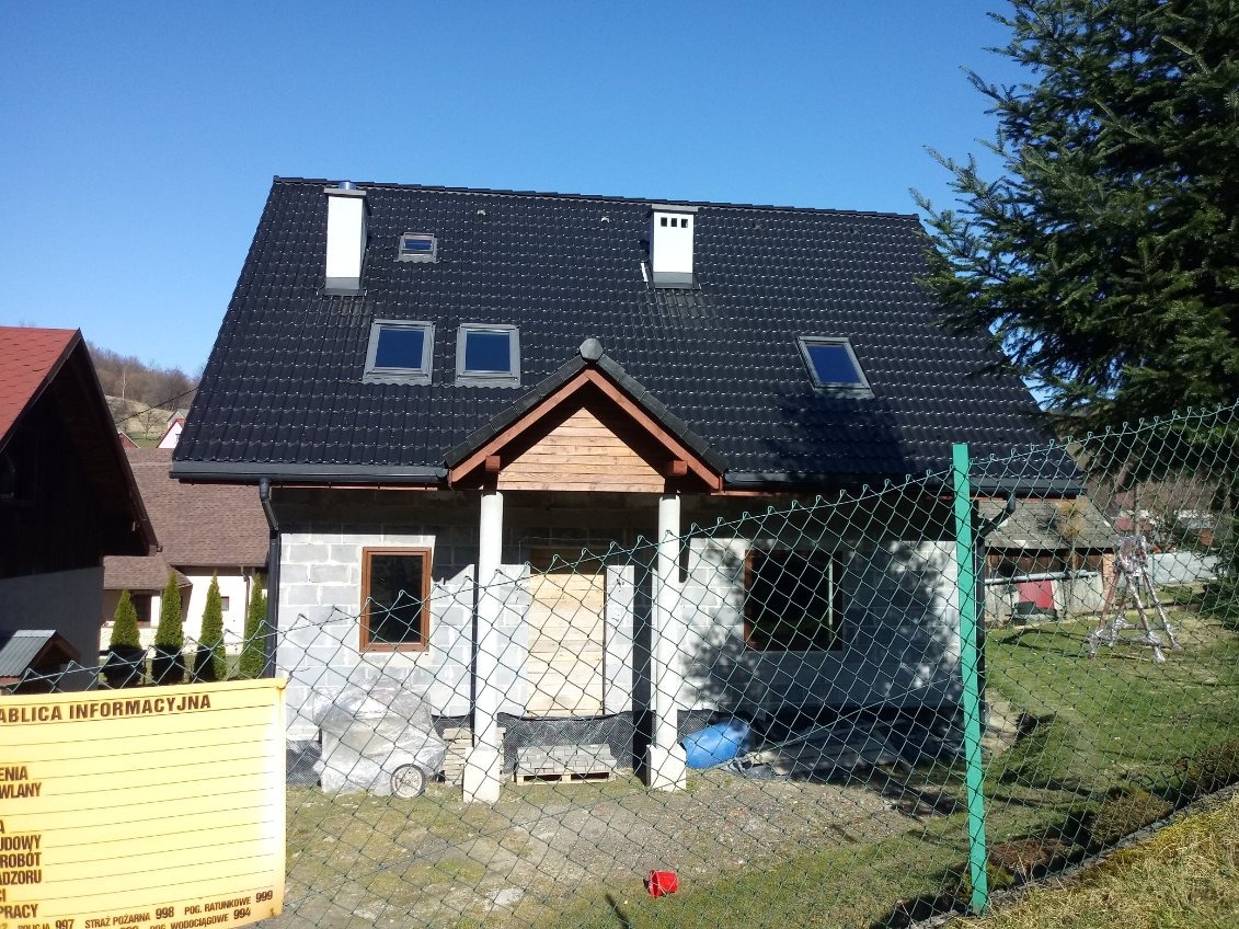 Surowy budynek z szarego bloczka, z ciemnym dachem z dachówką, widoczne okna dachowe, częściowo osłonięty zielonym ogrodzeniem z siatki, z tablicą informacyjną budowy po lewej stronie.