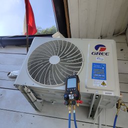 EKOZYSK Grzegorz Kotecki - Zewnętrzna jednostka klimatyzacji marki Gree zamontowana na białej, drewnianej ścianie budynku, podłączona do cyfrowego kolektora pomiarowego z niebieskimi przewodami, widoczna flaga Polski w tle.