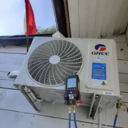 Zewnętrzna jednostka klimatyzacji marki Gree zamontowana na białej, drewnianej ścianie budynku, podłączona do cyfrowego kolektora pomiarowego z niebieskimi przewodami, widoczna flaga Polski w tle.