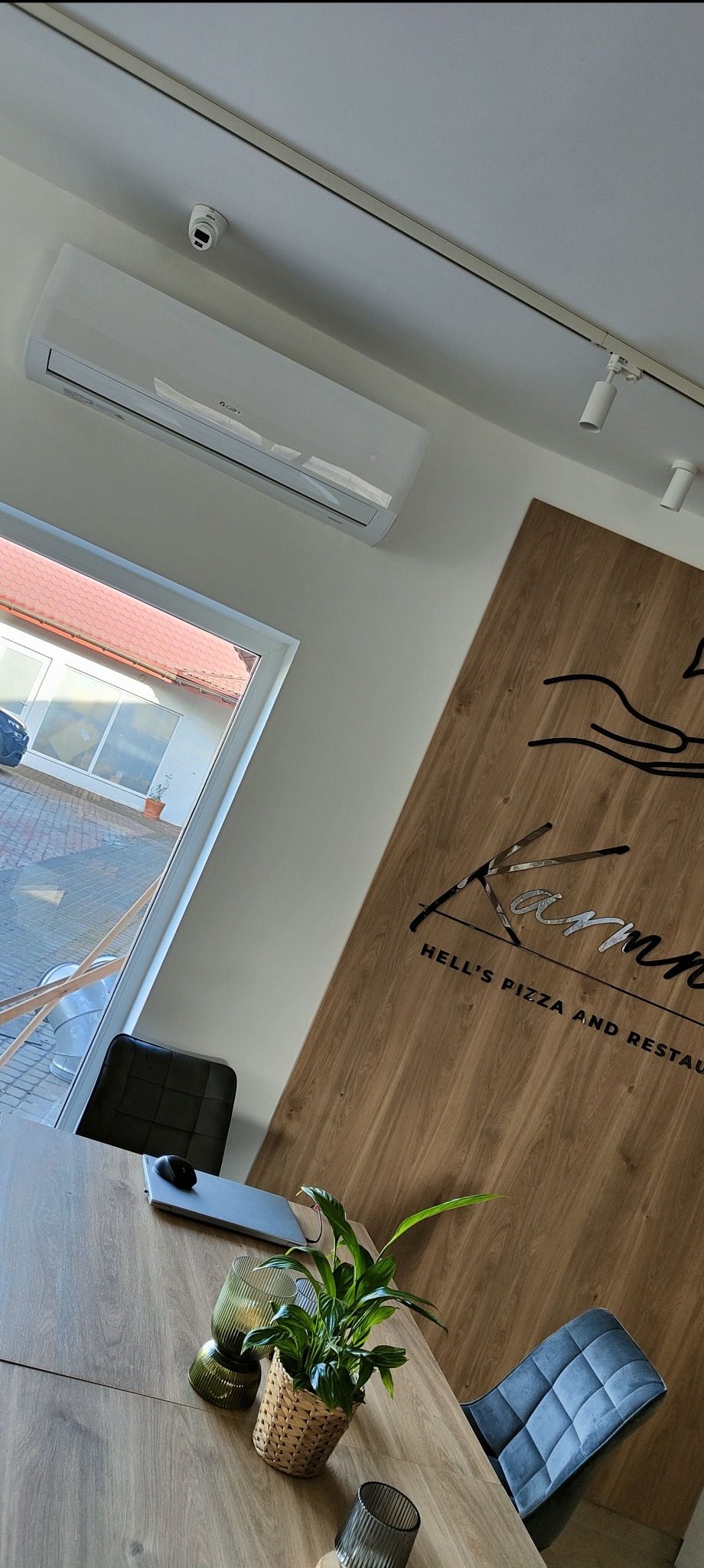 Wnętrze restauracji z zamontowanym klimatyzatorem ściennym marki Gree, widocznym nad oknem, obok lamp punktowych i dekoracyjnej drewnianej ściany z logo 'Hell's Pizza and Restaurant'. Na stole...