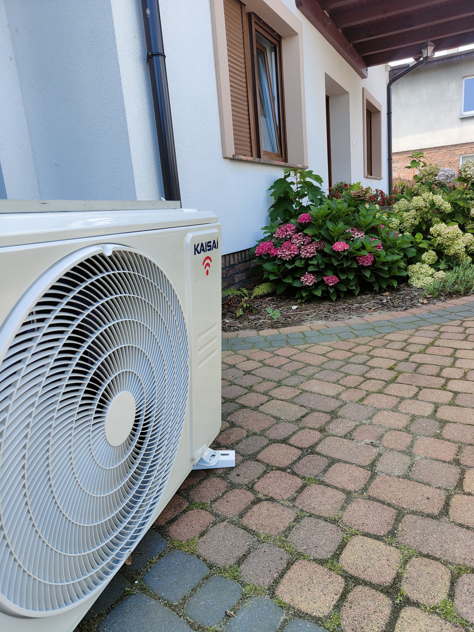 Zewnętrzna jednostka klimatyzacji Kaisai zamontowana przy ścianie budynku z brukowanym podjazdem i kwitnącymi hortensjami w tle.