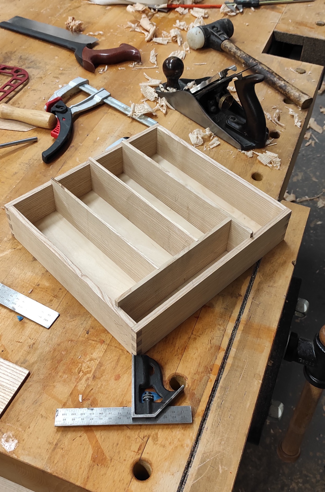 Drewniany organizer na narzędzia w trakcie tworzenia, widoczne dłuto, strug, młotek i ścisk stolarski na warsztacie z wiórami.