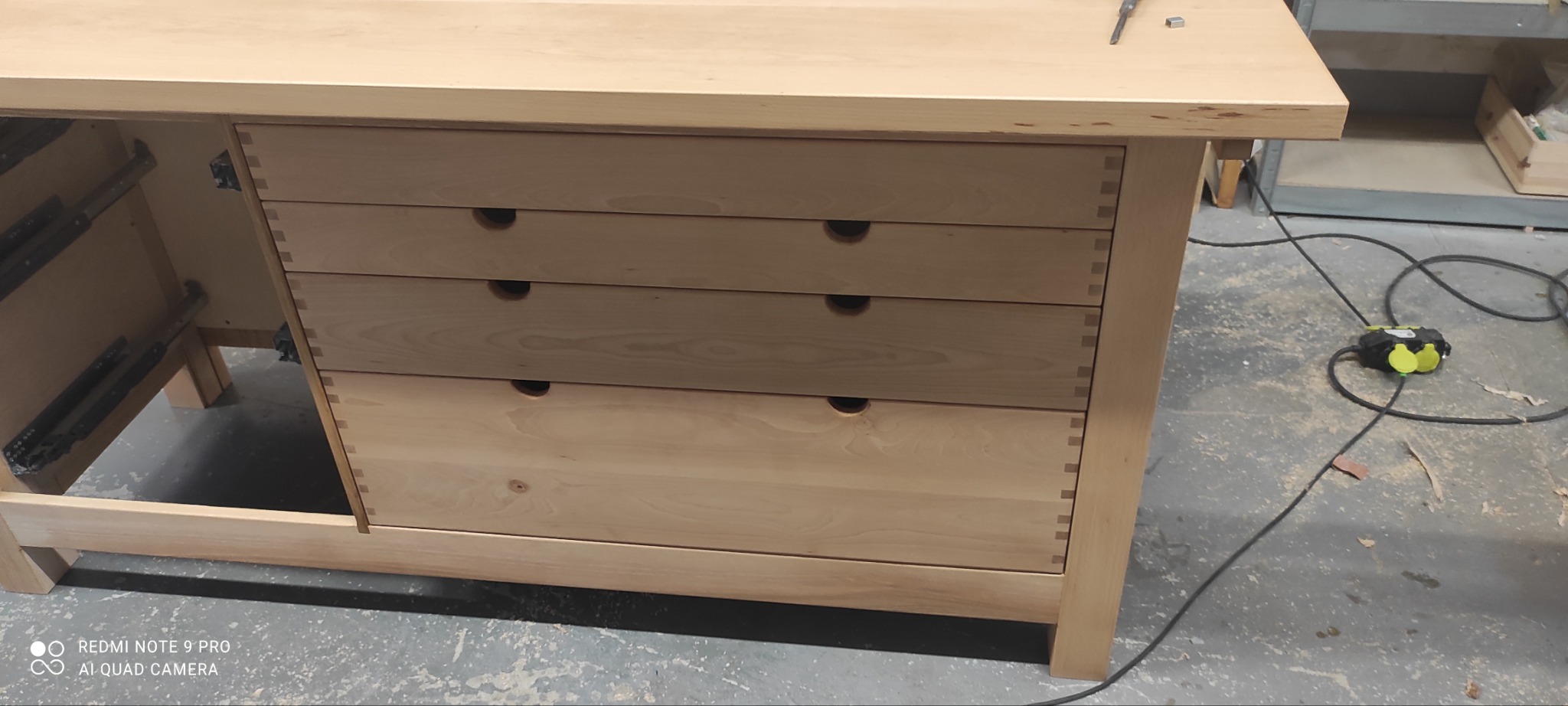 Niedokończona komoda z jasnego drewna z czterema szufladami, widoczne połączenia na jaskółczy ogon, w tle narzędzia i przedłużacz na podłodze warsztatu.