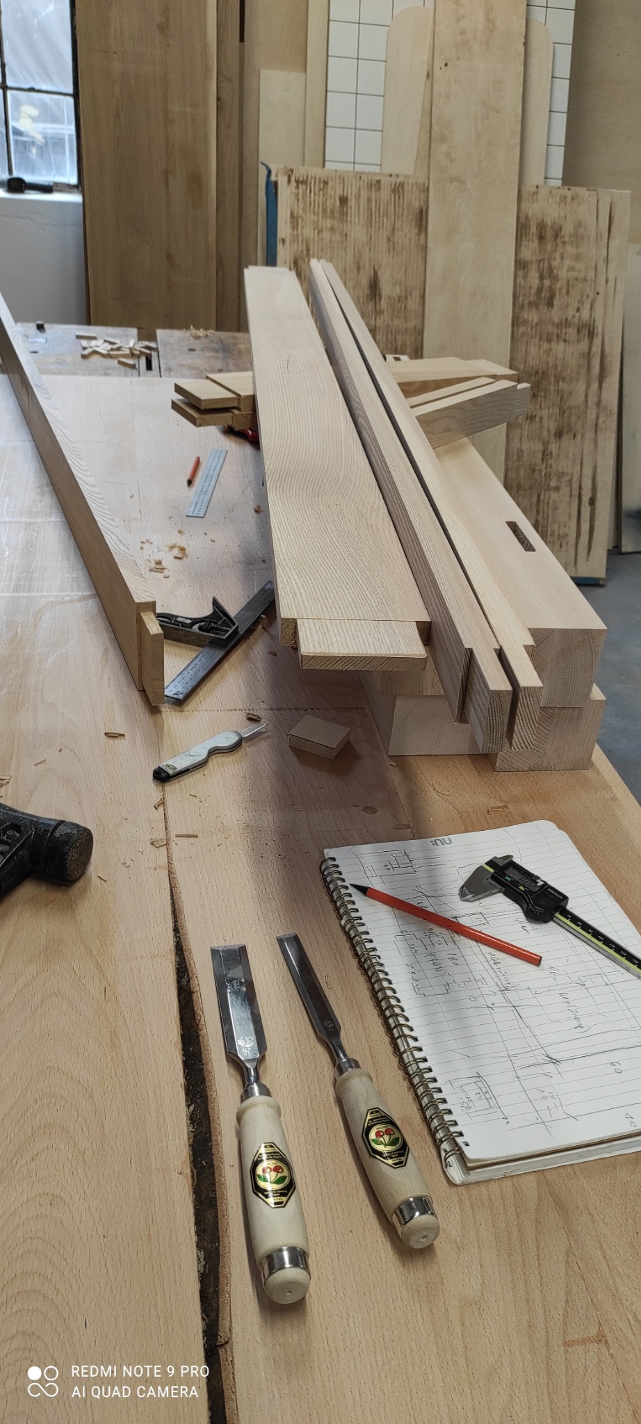 Warsztat stolarski: surowe drewniane elementy konstrukcyjne z wyfrezowanymi wpustami, dłuta z logo wiśni, notatnik z odręcznym szkicem, suwmiarka, kątownik stolarski i ołówek na drewnianym stole...