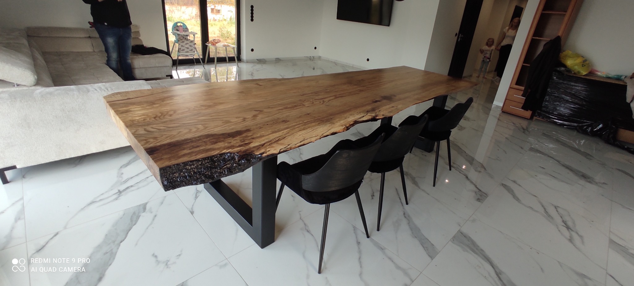 Stół z blatem z litego drewna o nieregularnych krawędziach, wsparty na metalowych nogach w kształcie litery U, otoczony czarnymi krzesłami w nowoczesnym wnętrzu z marmurową podłogą.