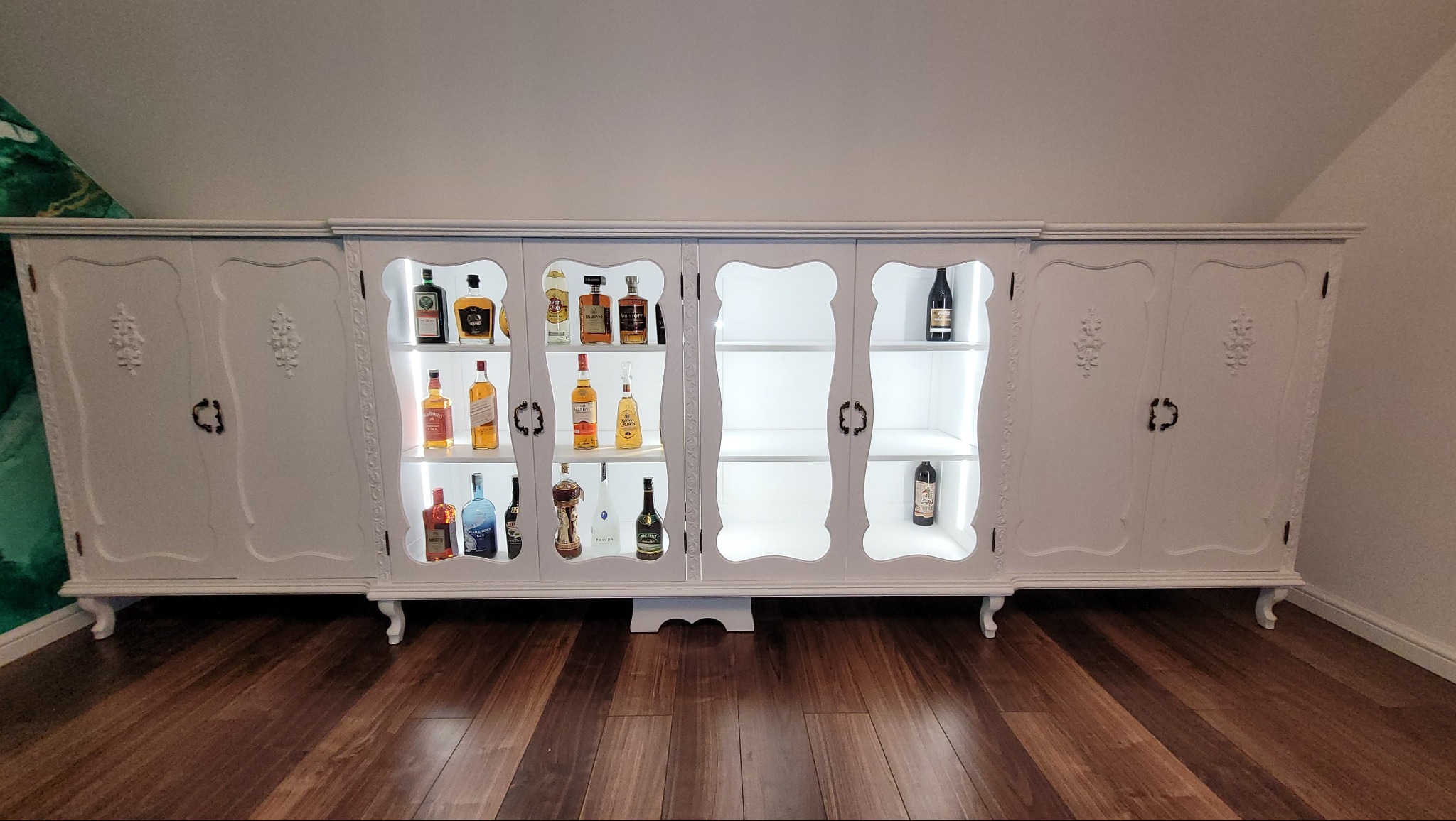 Biała, stylizowana szafa barowa z podświetlanymi półkami, wypełniona butelkami alkoholu. Dekoracyjne zdobienia i ciemne uchwyty na tle drewnianej podłogi.