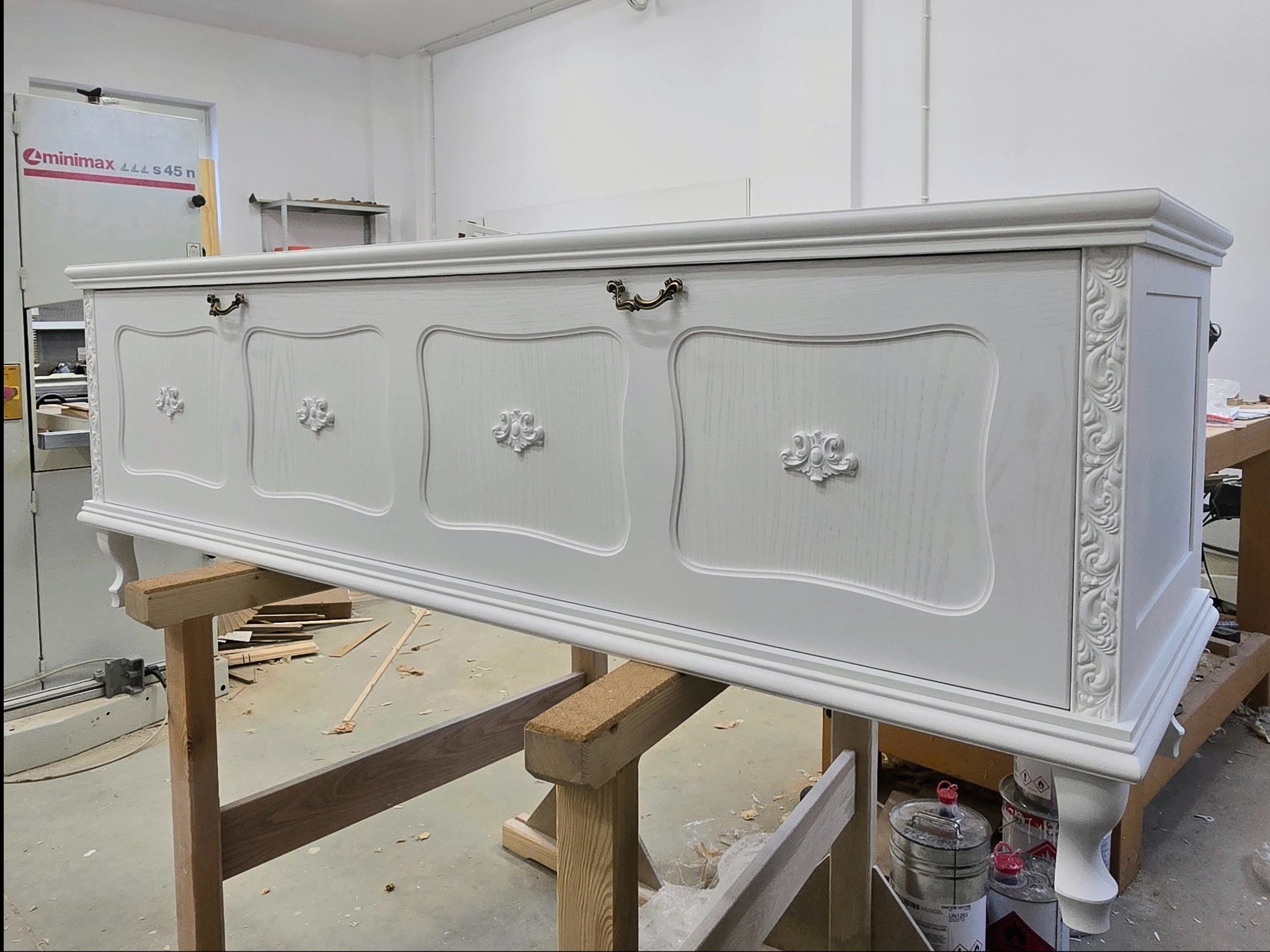 Biała, ręcznie robiona skrzynia z dekoracyjnymi uchwytami i ornamentami, ustawiona na drewnianych kozłach w warsztacie. Widoczne narzędzia i materiały.