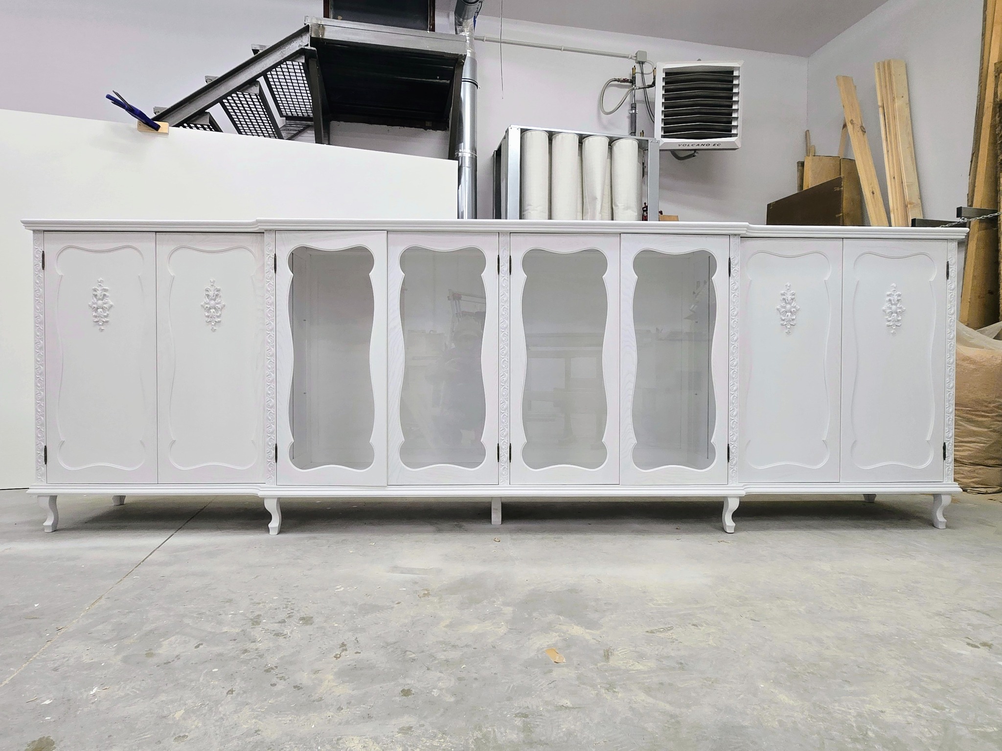 Biała, stylowa komoda z przeszklonymi drzwiczkami i dekoracyjnymi ornamentami, ustawiona w warsztacie na tle betonowej podłogi i instalacji wentylacyjnej.