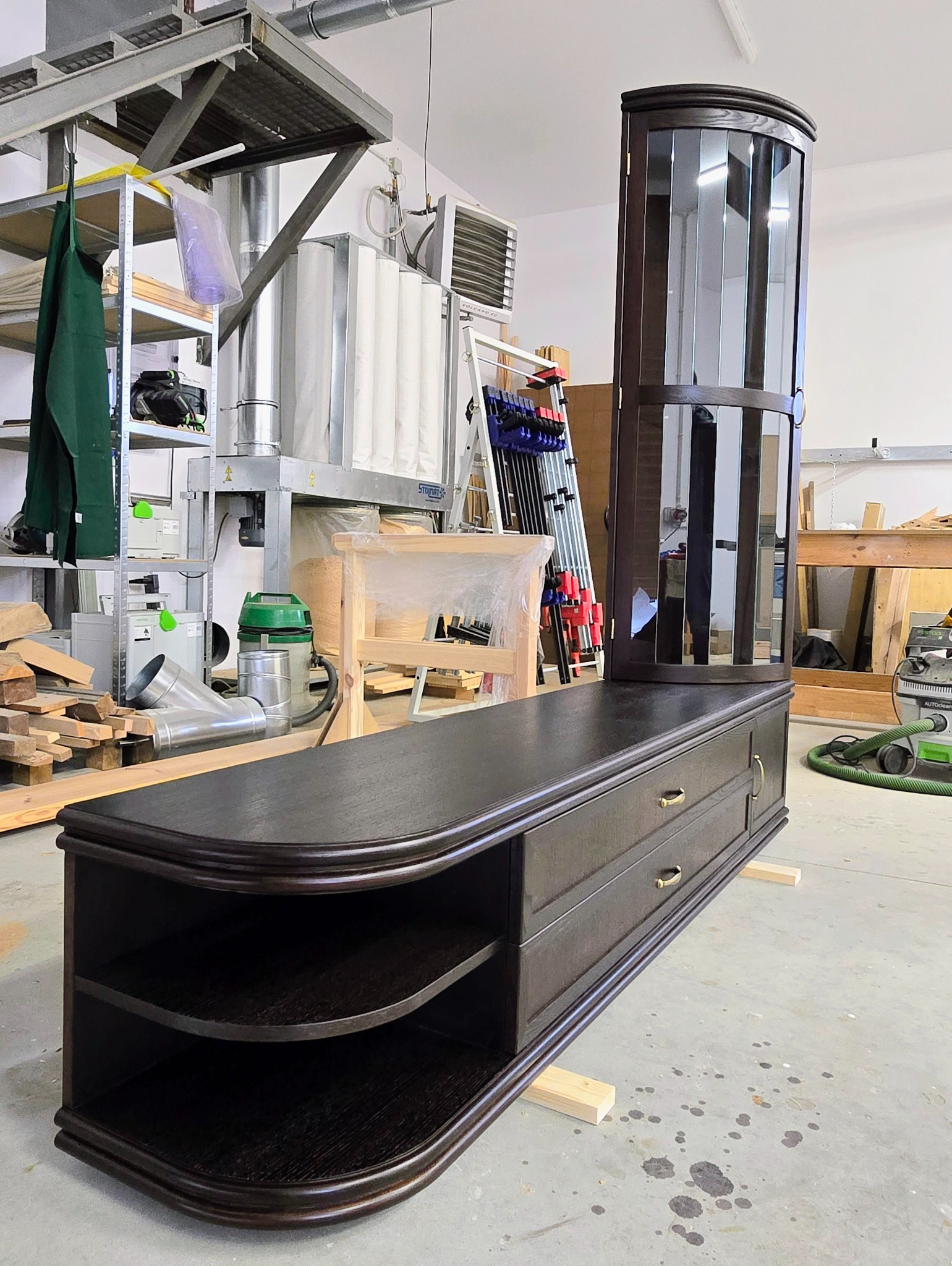 Ciemna, fornirowana szafka RTV z szufladami i półkami, oraz witryna w tle, w warsztacie stolarskim. Widoczne narzędzia i maszyny stolarskie.