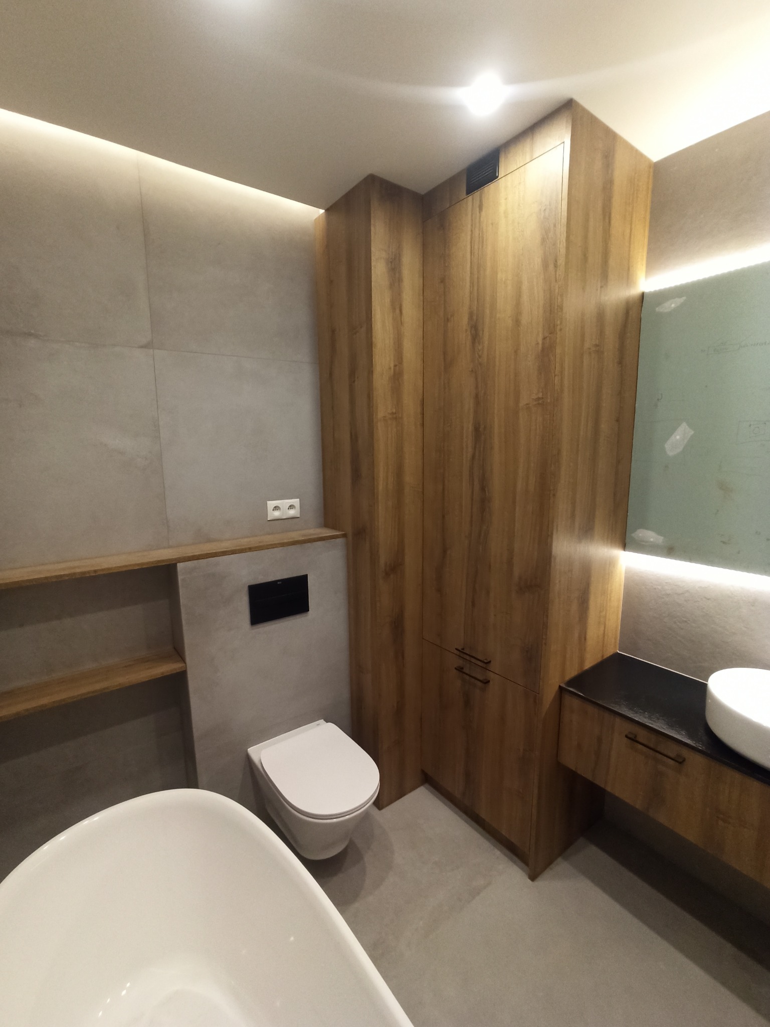 Nowoczesna łazienka z wanną wolnostojącą, podwieszaną toaletą i szafką z umywalką nablatową, wykończona płytkami w odcieniach szarości i drewnopodobnymi panelami na szafkach i półkach, oświetlona...