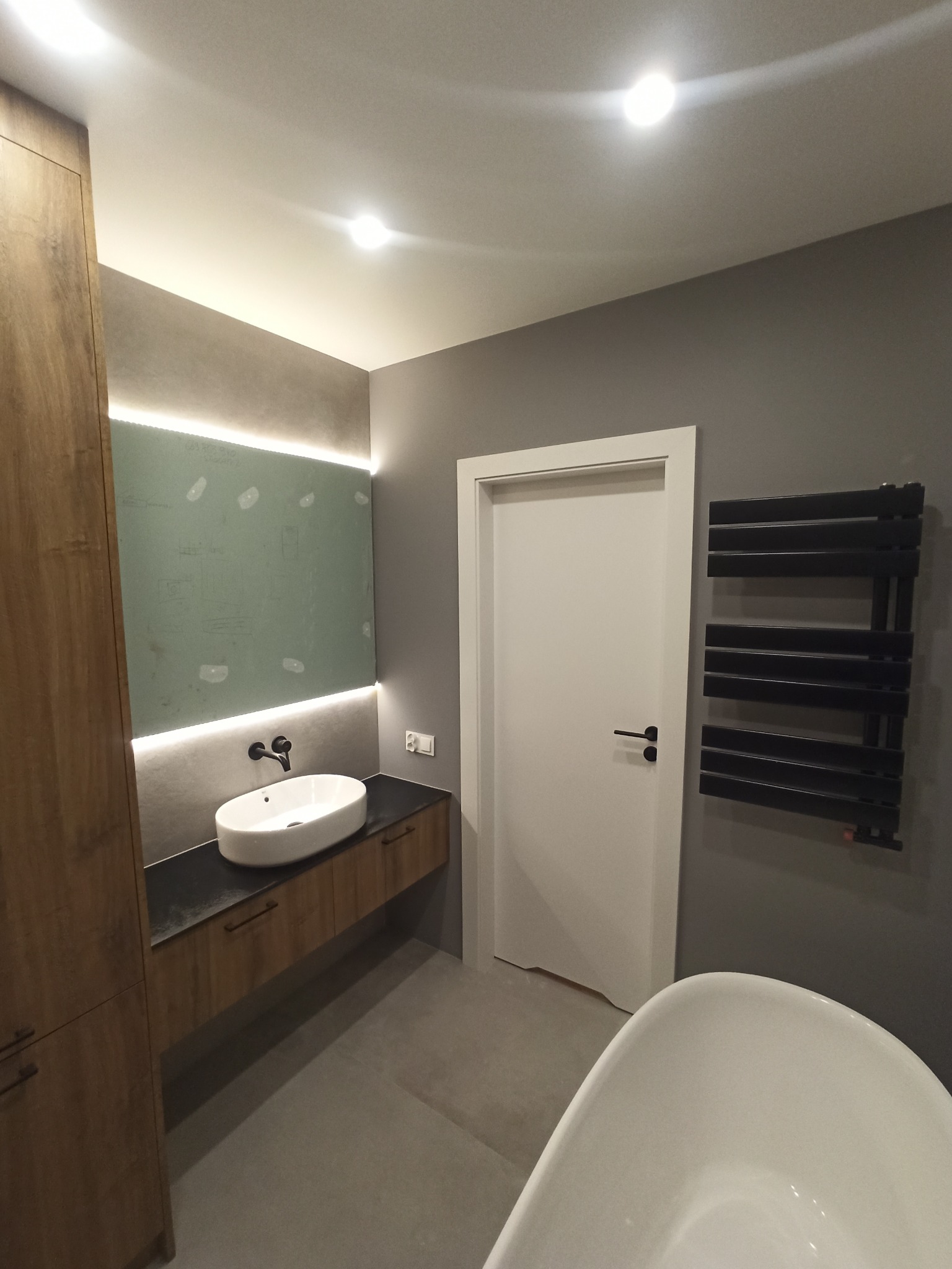 Nowoczesna łazienka z owalną umywalką na drewnopodobnej szafce z czarnym blatem, podświetlonym lustrem, grzejnikiem drabinkowym i częściowo widoczną białą wanną wolnostojącą.