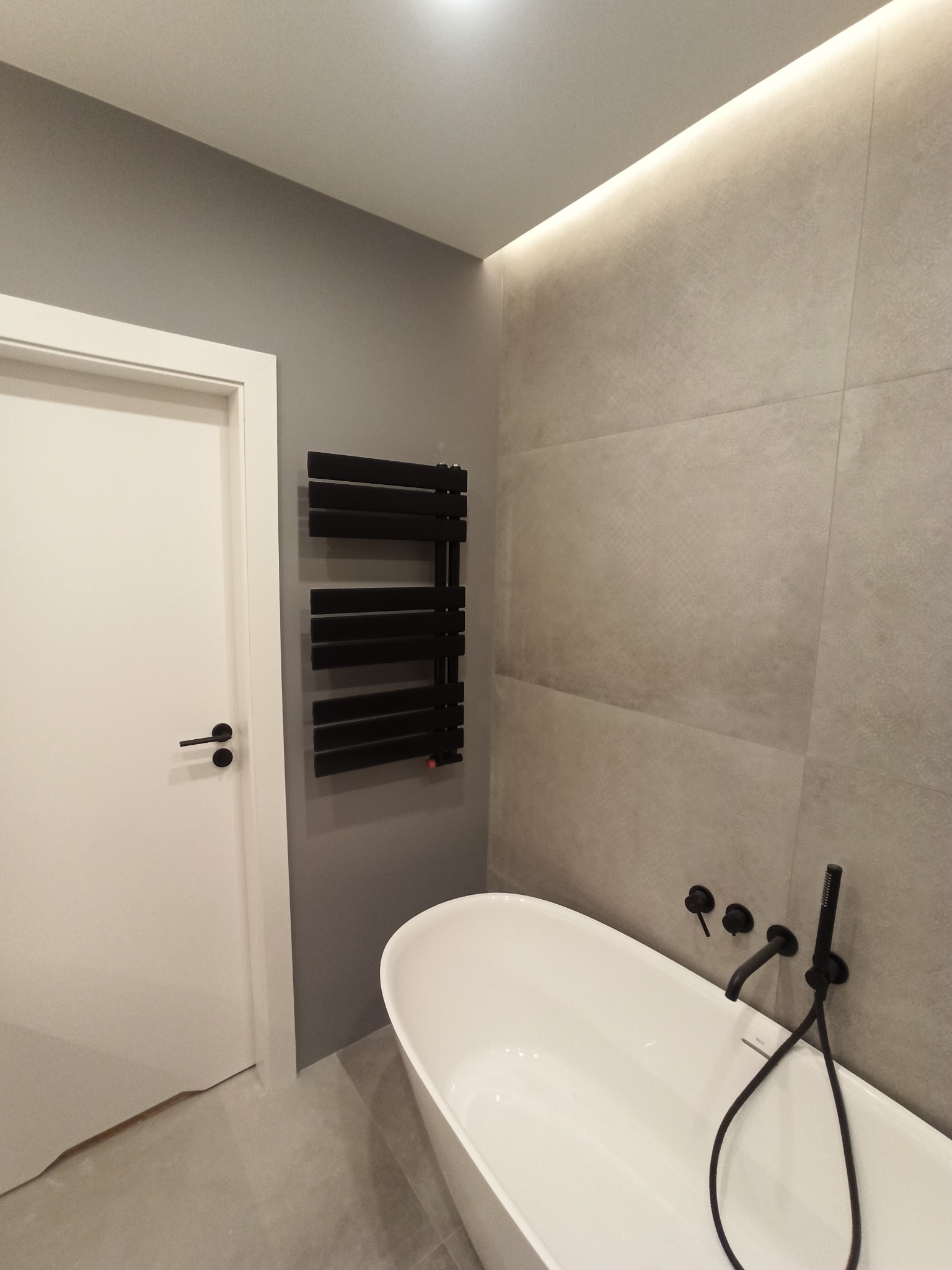 Nowoczesna łazienka z wolnostojącą białą wanną, czarną armaturą ścienną i grzejnikiem drabinkowym w kolorze czarnym, szara ściana z płytek, białe drzwi z czarną klamką oraz oświetlenie LED.