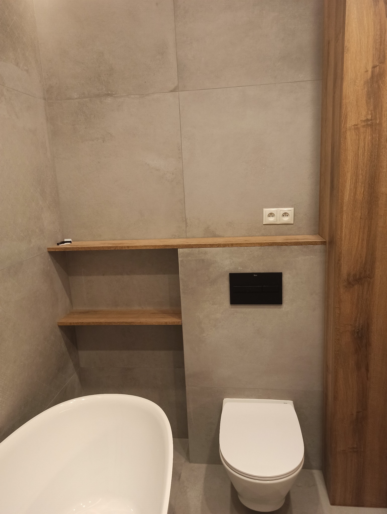 Nowoczesna łazienka z szarymi płytkami, wiszącą toaletą z czarnym przyciskiem spłukującym, wolnostojącą wanną i drewnianymi półkami wnękowymi.