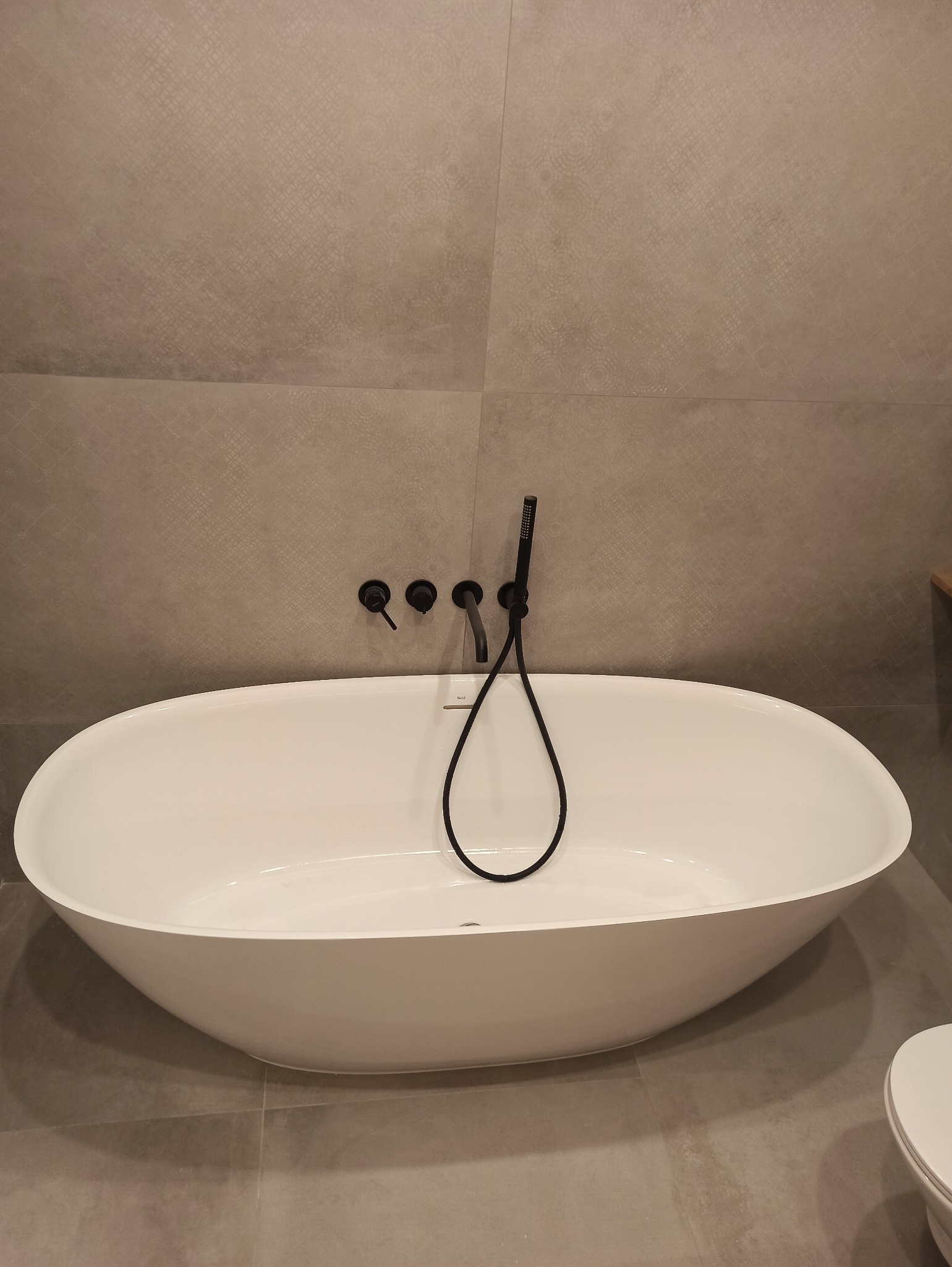 Nowoczesna, wolnostojąca wanna owalna z czarną armaturą ścienną na tle płytek w odcieniach szarości z delikatnym wzorem.