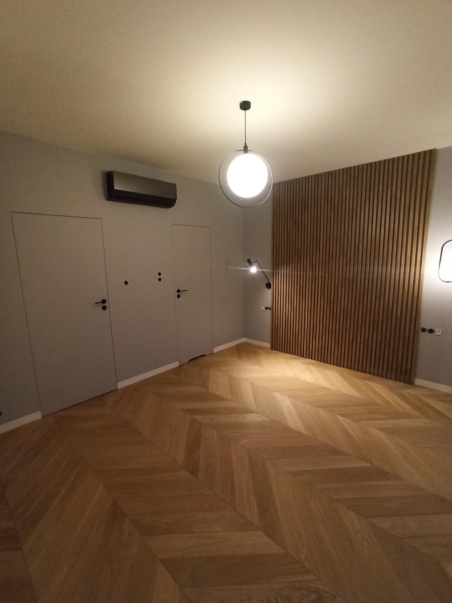 Nowoczesne wnętrze z drewnianą podłogą ułożoną w jodełkę, drewnianą ścianą z pionowych listew, ukrytymi drzwiami i minimalistycznym oświetleniem, w tym lampą wiszącą z okrągłym kloszem i kinkietem.