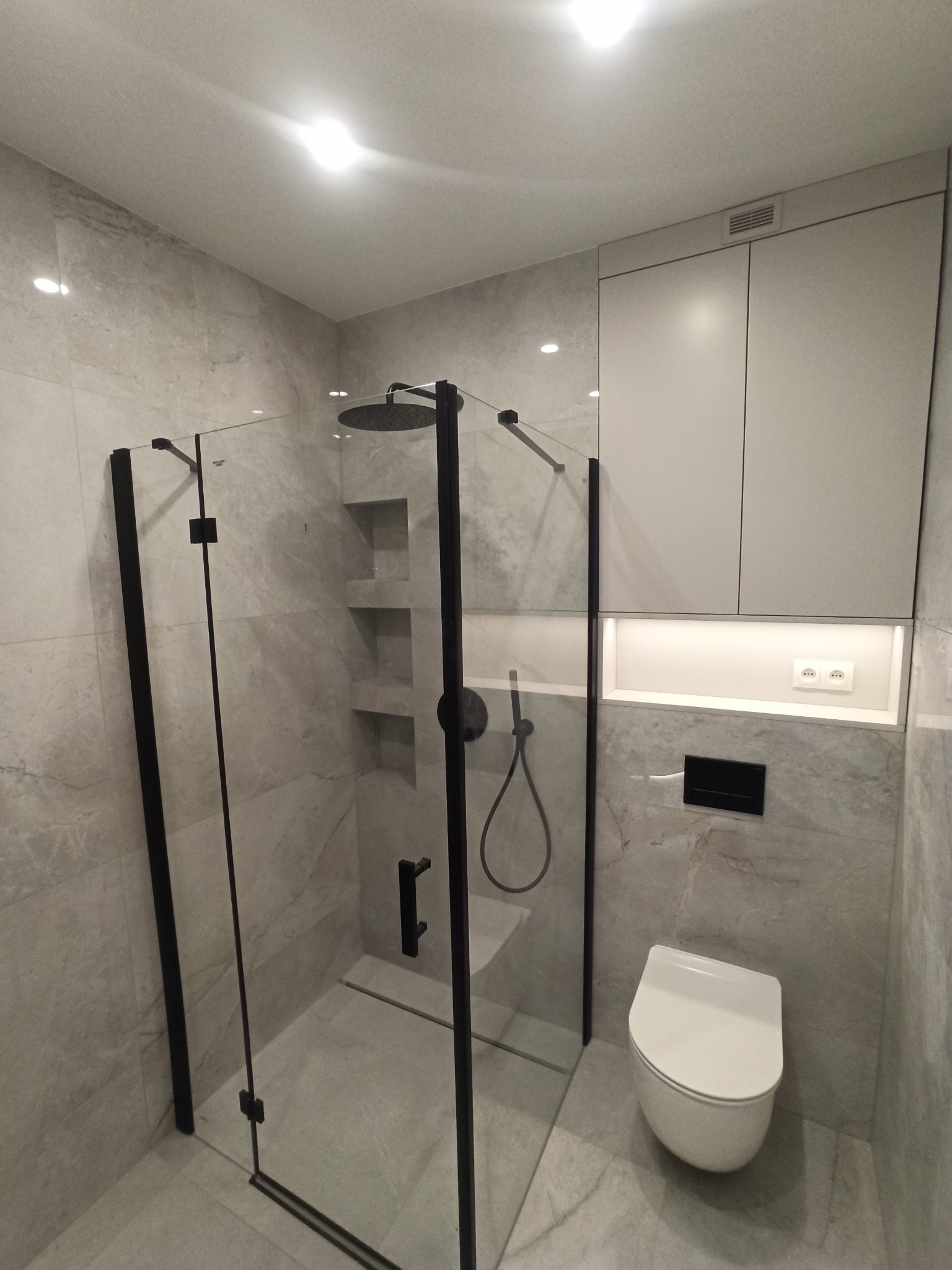 Nowoczesna łazienka w odcieniach szarości z kabiną prysznicową typu walk-in z czarnymi profilami, wiszącą toaletą, szafką i wnękami w ścianie wyłożonymi płytkami imitującymi marmur.
