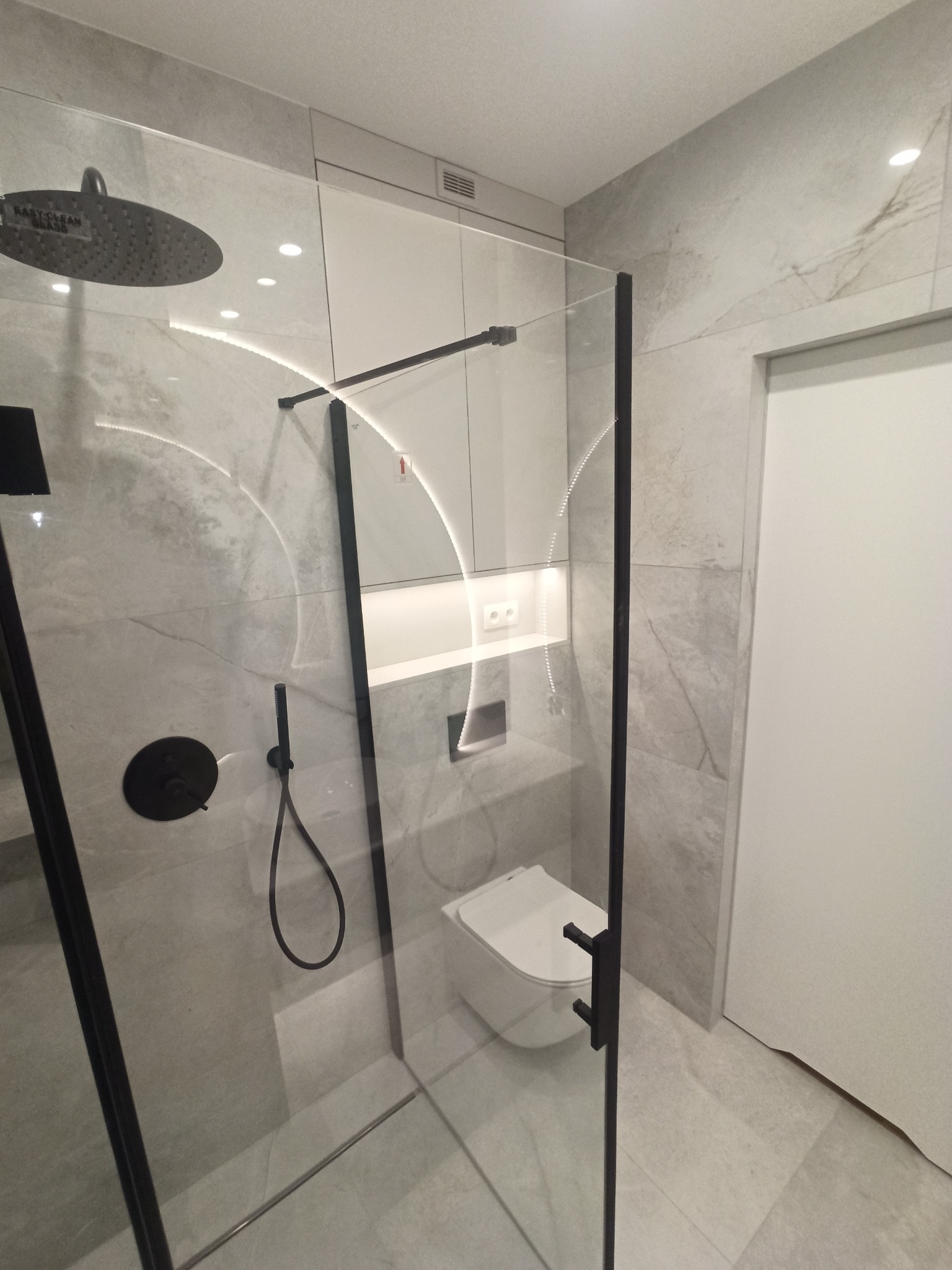 Nowoczesna kabina prysznicowa z czarnymi elementami, przeszklona, z deszczownicą i minimalistyczną armaturą na tle płytek imitujących marmur. W tle widoczna podświetlana wnęka z toaletą podwieszaną.