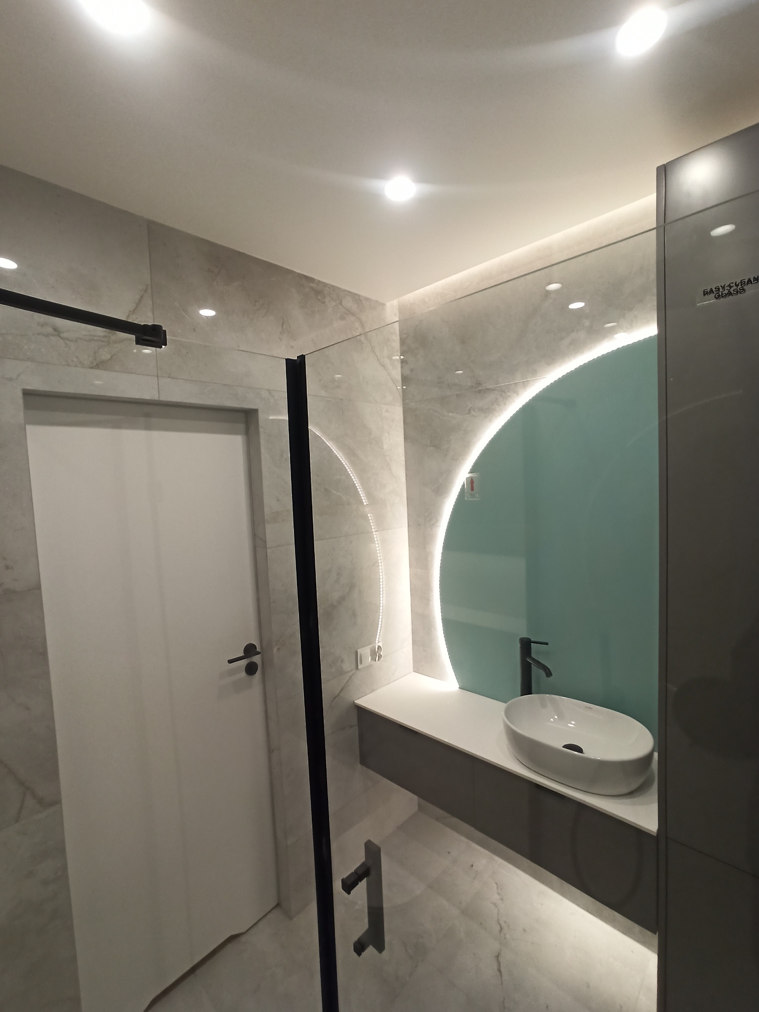 Nowoczesna łazienka z owalną umywalką nablatową, podświetlanym lustrem w kształcie półksiężyca, kabiną prysznicową z czarnymi profilami i drzwiami oraz płytkami imitującymi marmur