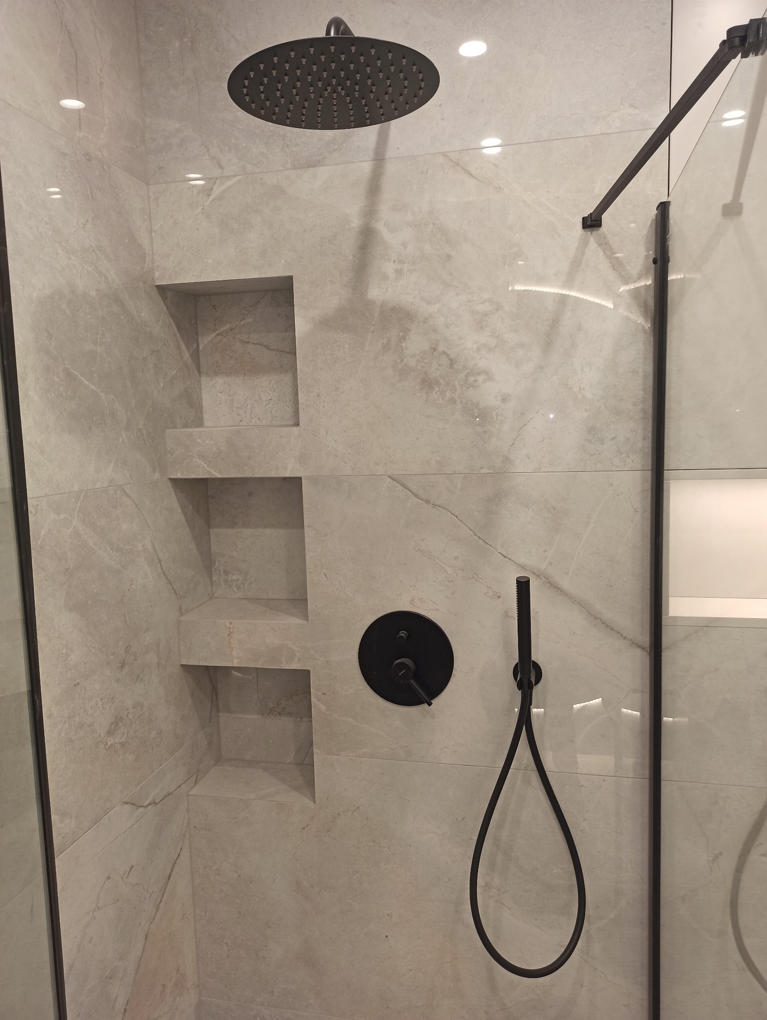 Nowoczesna kabina prysznicowa z deszczownicą i czarną armaturą na marmurowych płytkach, wbudowane półki w ścianie.
