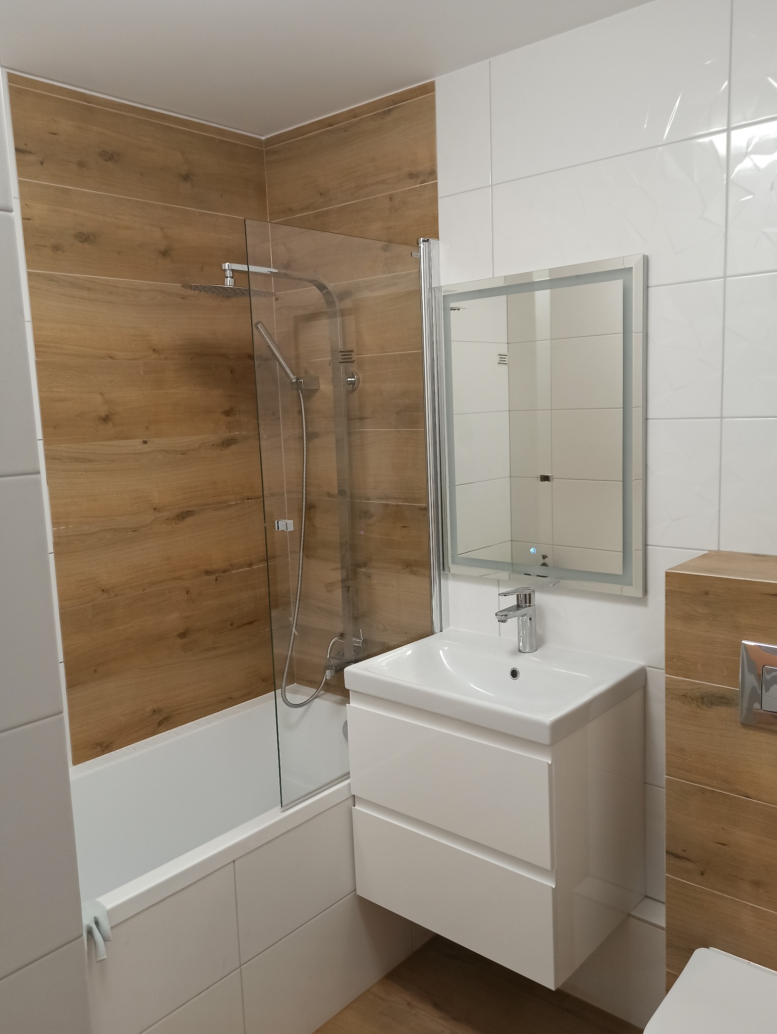 Nowoczesna łazienka z drewnopodobnymi i białymi płytkami, kabiną prysznicową za szklanym ekranem, białą umywalką z szafką i podświetlanym lustrem.