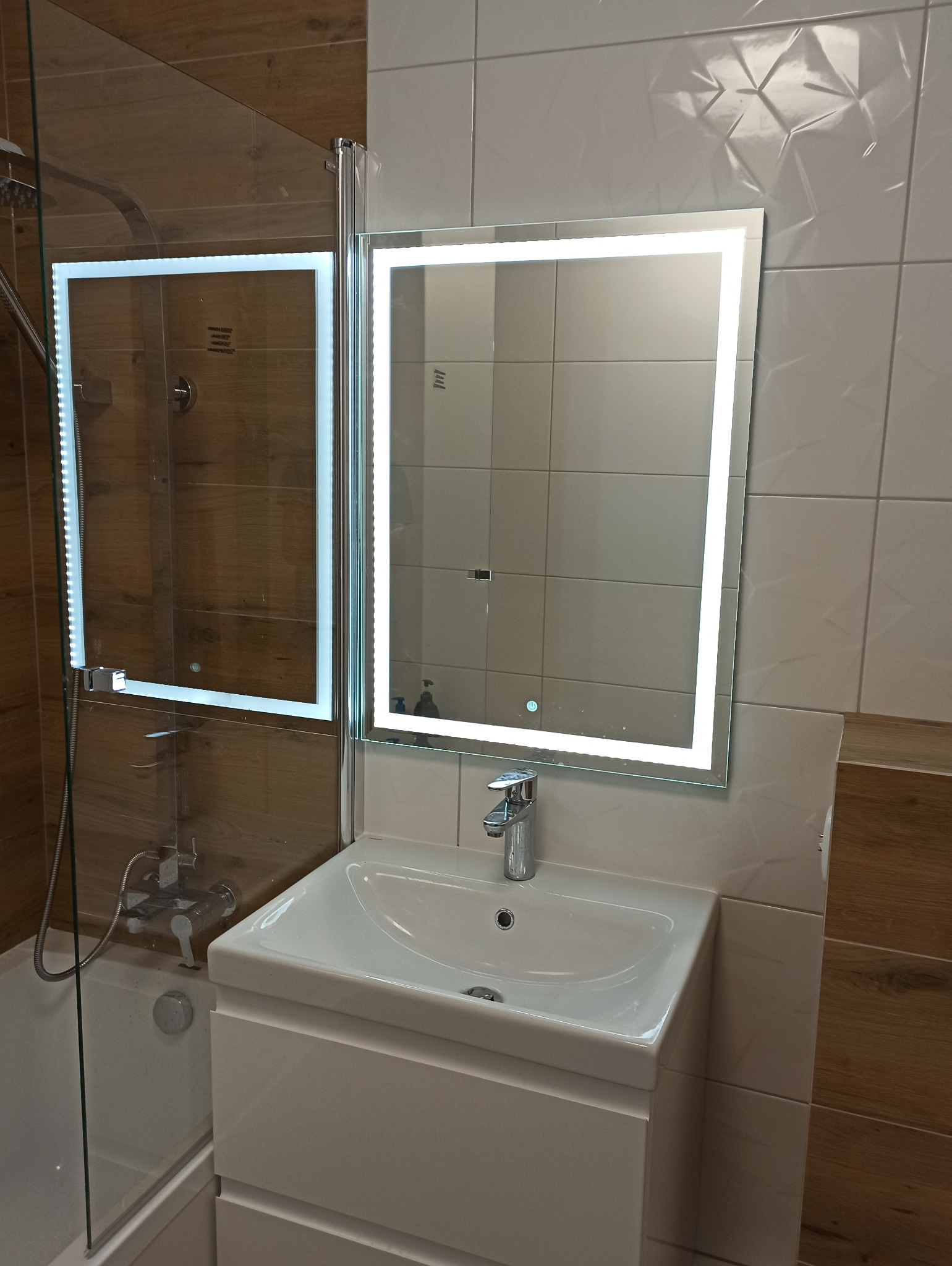 Nowoczesna łazienka z podświetlanym lustrem, umywalką z szafką w kolorze białym, kabiną prysznicową ze szklaną ścianką i brązowymi płytkami imitującymi drewno.