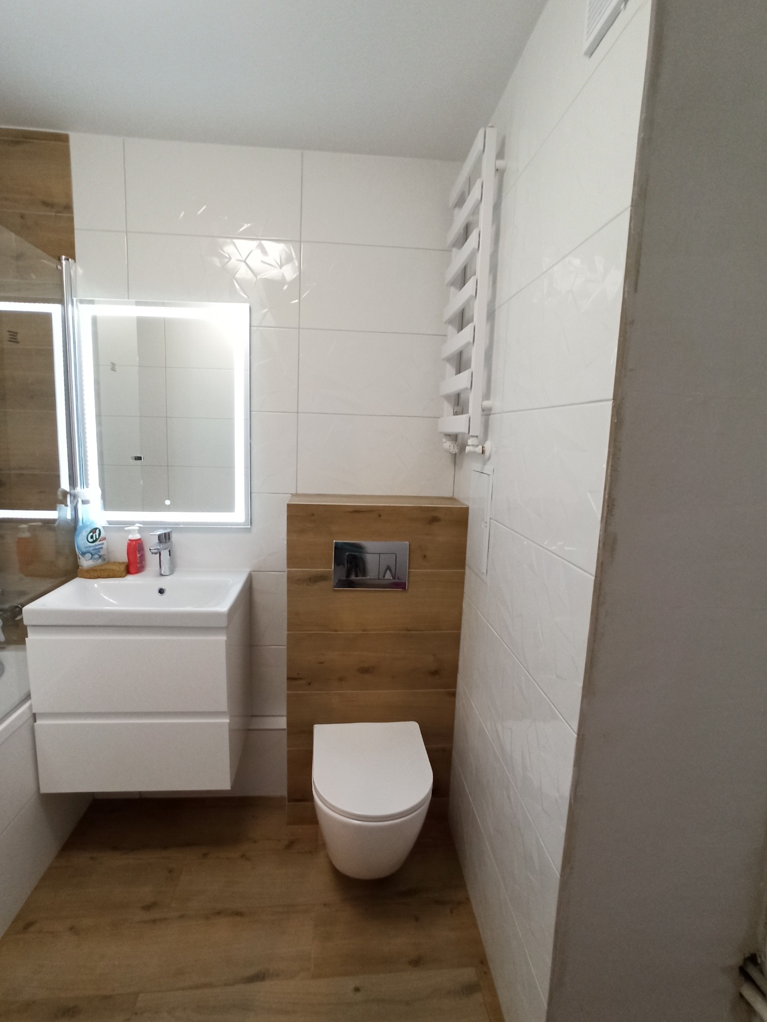 Nowoczesna łazienka z białymi płytkami, drewnopodobną zabudową geberit, podświetlanym lustrem nad białą szafką z umywalką i grzejnikiem drabinkowym.