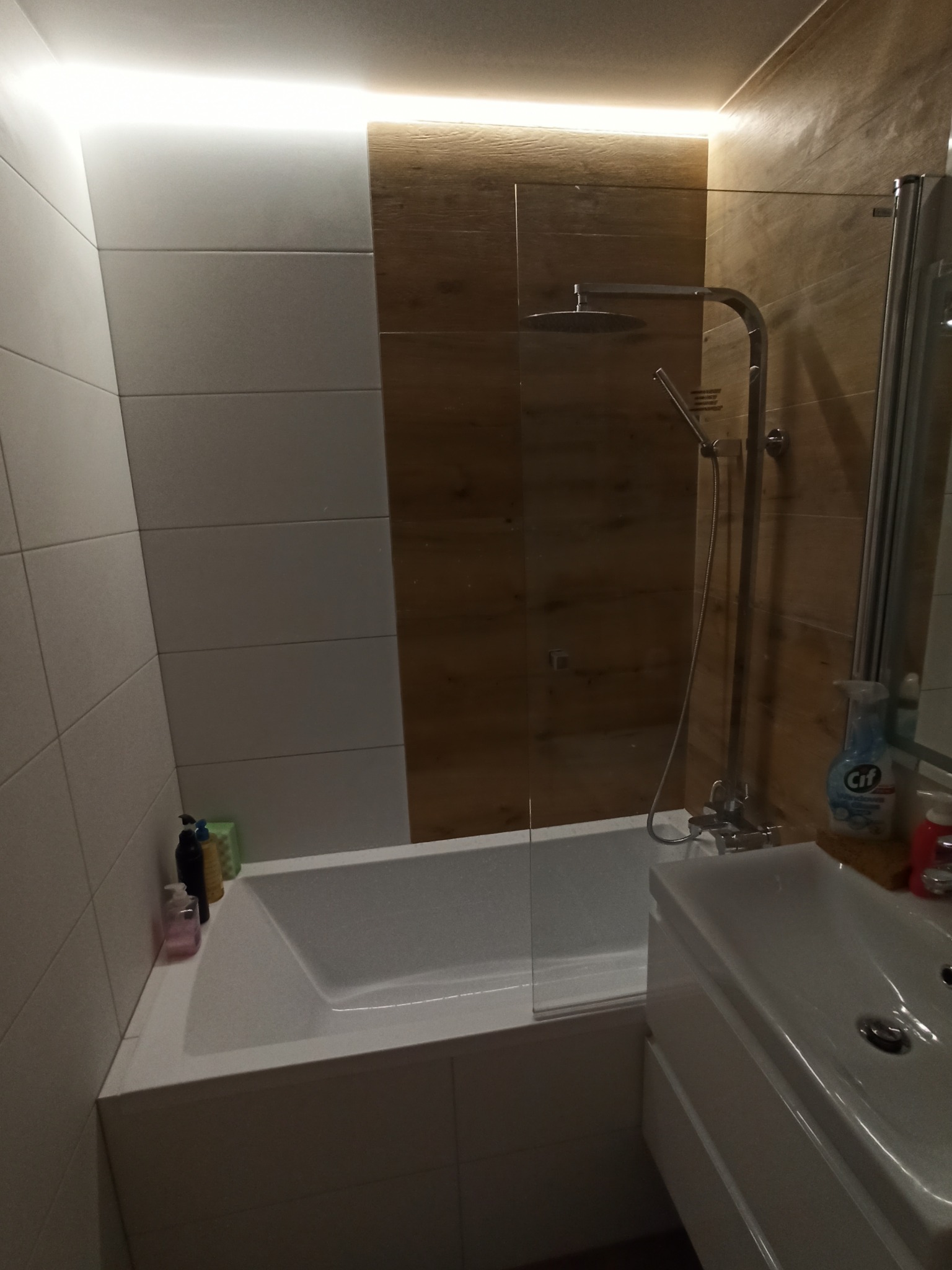 Nowoczesna łazienka z białą wanną, szklanym parawanem nawannowym i drewnopodobnymi płytkami, oświetlona subtelnym światłem LED pod sufitem.