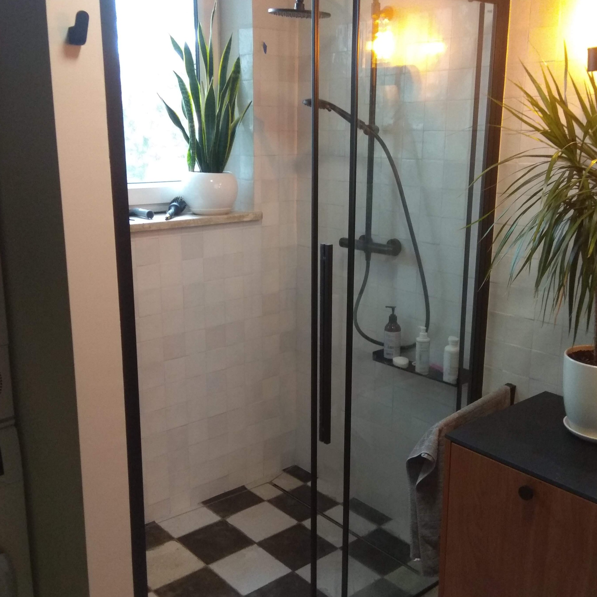 Nowoczesna kabina prysznicowa z czarnymi profilami, białe płytki ścienne, podłoga w szachownicę, rośliny doniczkowe i akcesoria łazienkowe na półce.