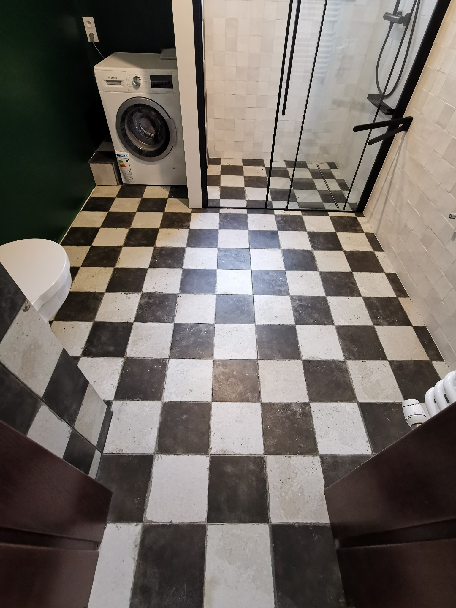 Szachownica z płytek w łazience z kabiną prysznicową typu walk-in i pralką, widok zza progu.