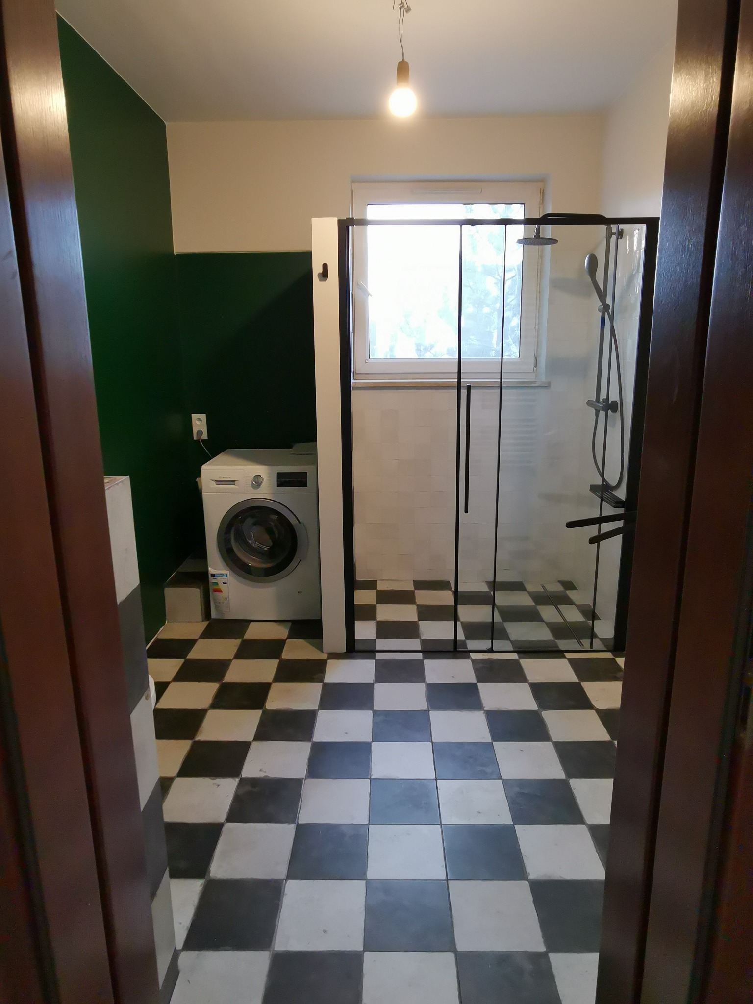 Ukończona łazienka z kabiną prysznicową typu walk-in z czarnymi profilami, pralką Bosch oraz podłogą w szachownicę, widok zza otwartych drzwi.