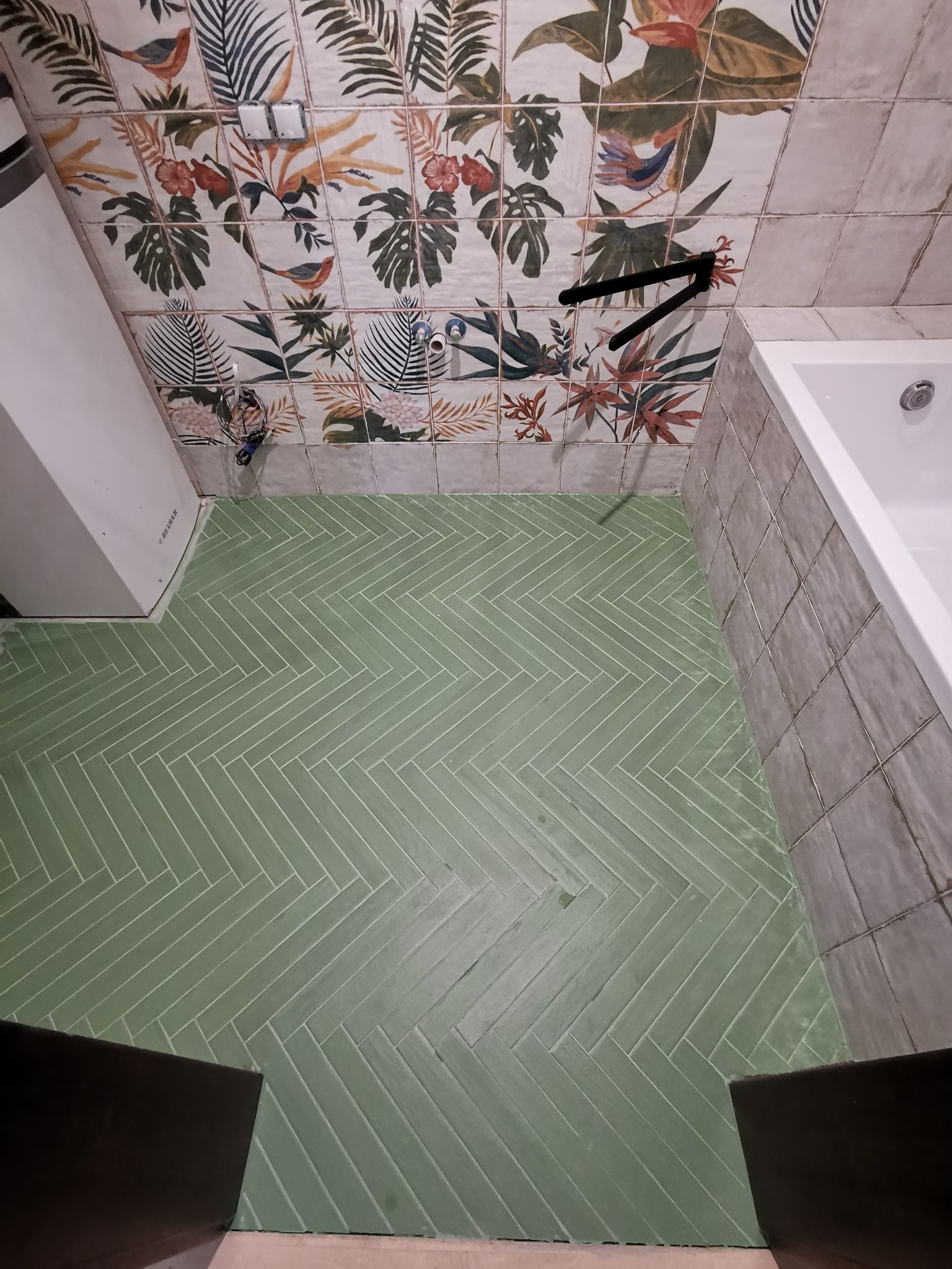Jasnozielona podłoga w łazience wyłożona płytkami w jodełkę, ściany z dekoracyjnymi płytkami w motywy roślinne i ptasie, fragment wanny.