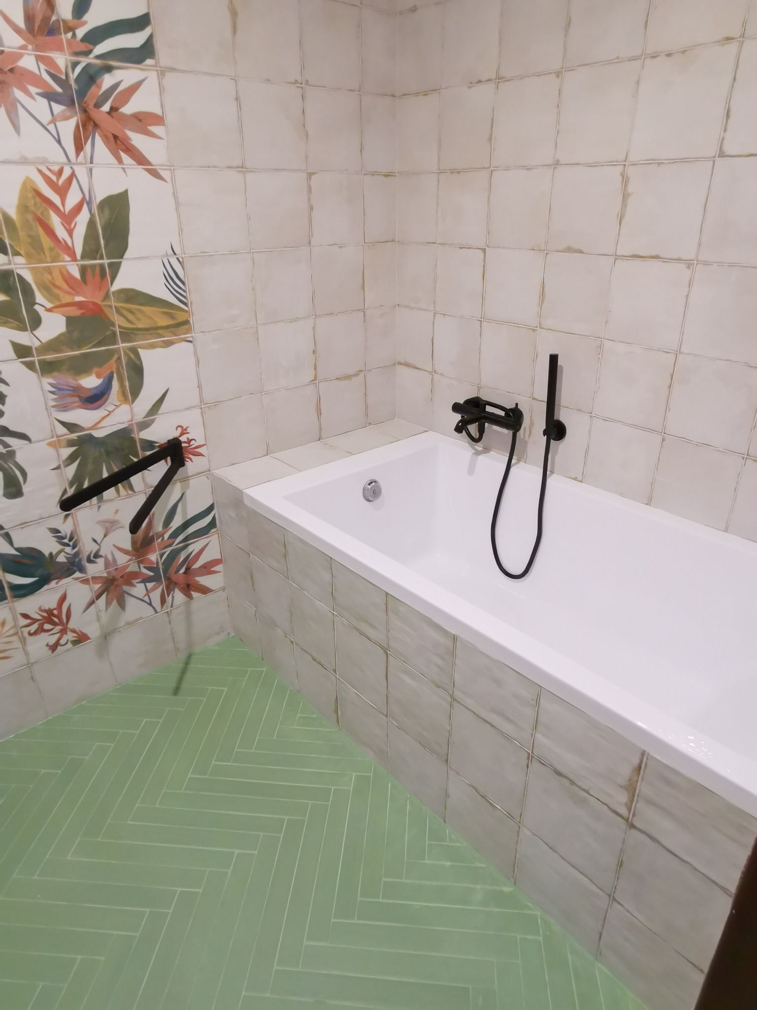 Nowoczesna łazienka z białą wanną, czarną armaturą i zieloną podłogą ułożoną w jodełkę, ściany wyłożone kwadratowymi płytkami z motywem roślinnym.